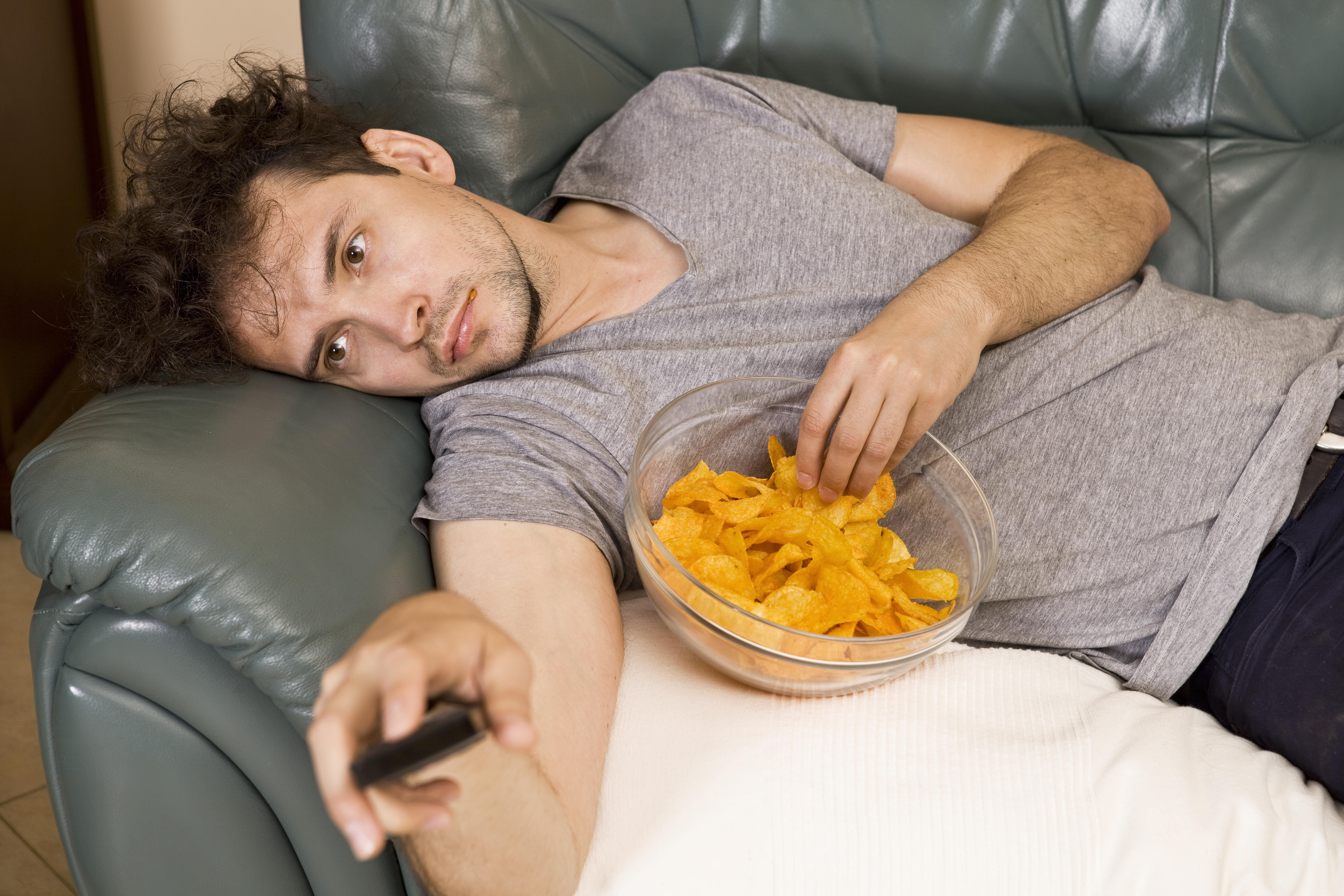 A lazy man laying on a couch watching television while eating snacks | Source: Getty Images