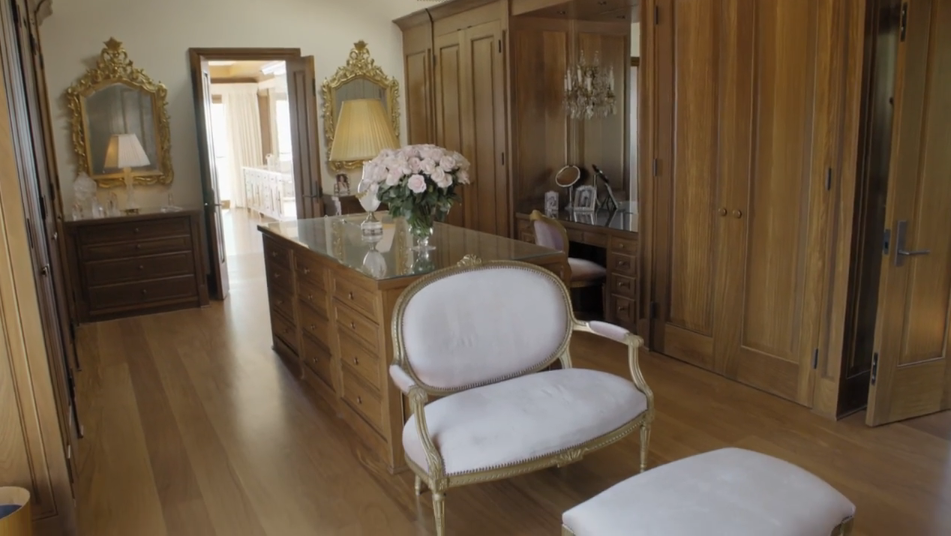 A glass-topped central island with built-in drawers divides this spacious walk-in closet, complemented by a plush, gold-trimmed settee and matching ottoman in the foreground. Floor-to-ceiling wooden wardrobes line the walls, while a vanity desk with a mirrored alcove and gilded wall mirrors add a touch of classic elegance. | Source: YouTube/Architectural Digest