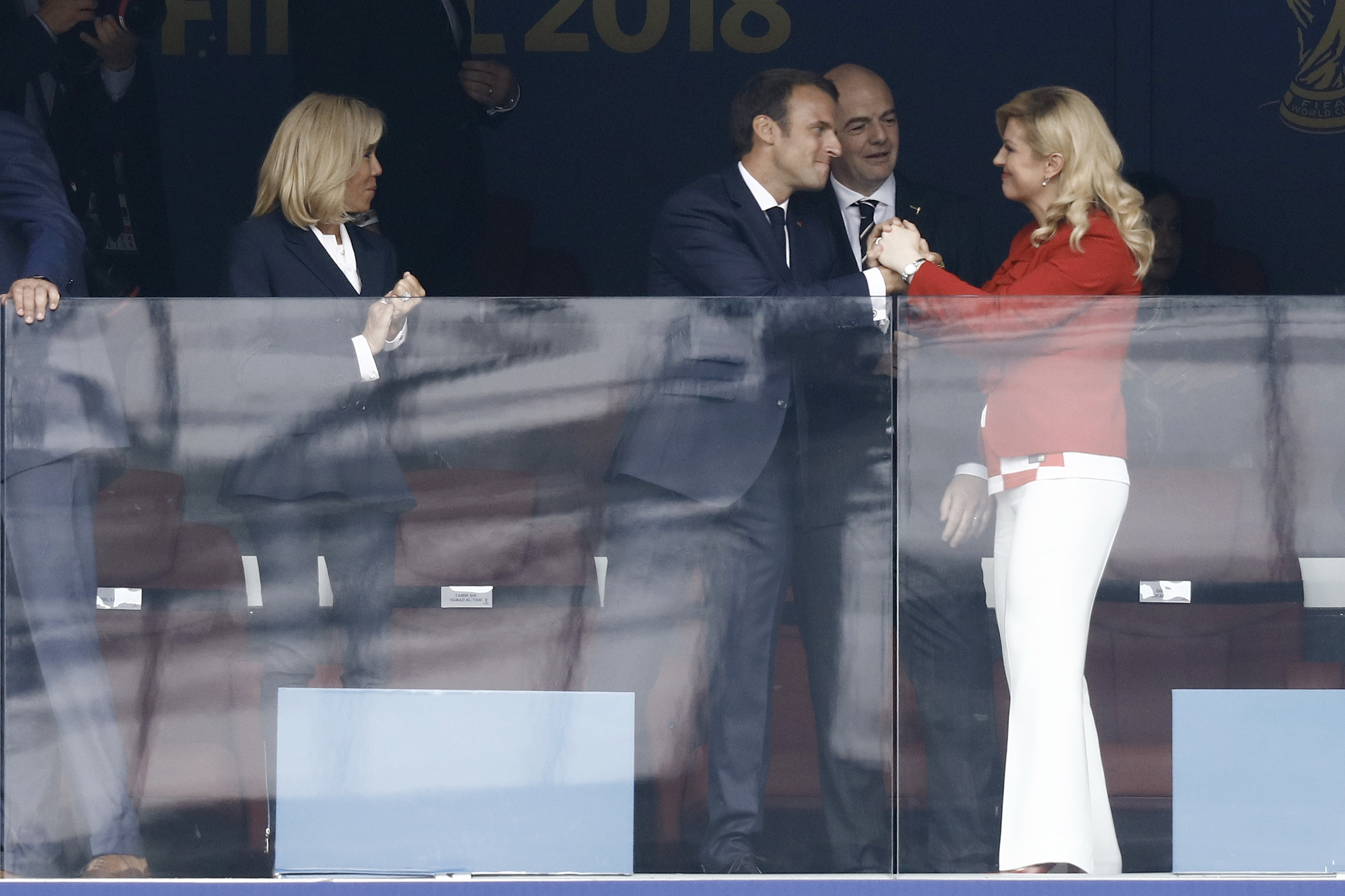 Emmanuel Macron is seen warmly greeting Kolinda Grabar-Kitarović with a firm handshake while smiling, as Brigitte Macron looks on from the side. The interaction takes place in a viewing box during the 2018 FIFA World Cup final.