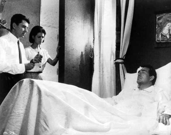 Philippe Nicaud et Claudia Cardinale debout au lit d'Ugo Tognazzi dans une scène du film "Le magnifique cocu", 1964. | Photo : Getty Images