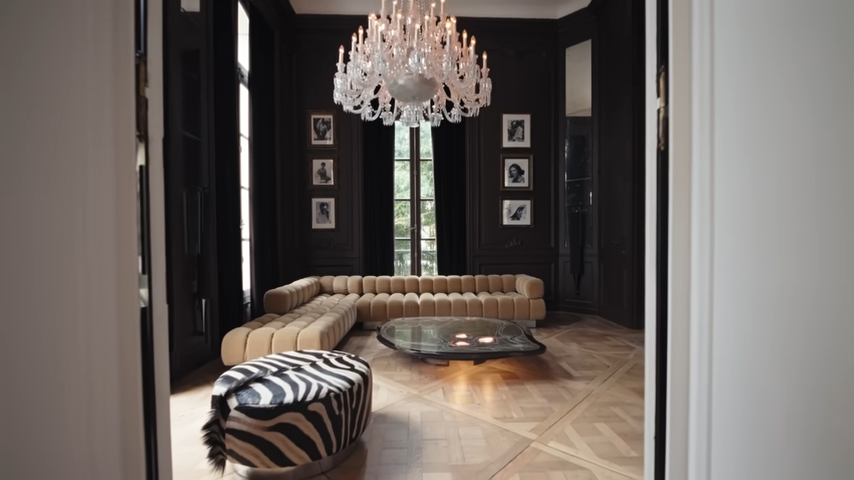 A wide view of the moody "Roxie Room" featuring a plush tan modular sofa, a custom organic coffee table with glowing agate slices, and a zebra-print ottoman under a crystal chandelier. | Source: YouTube/@Archdigest