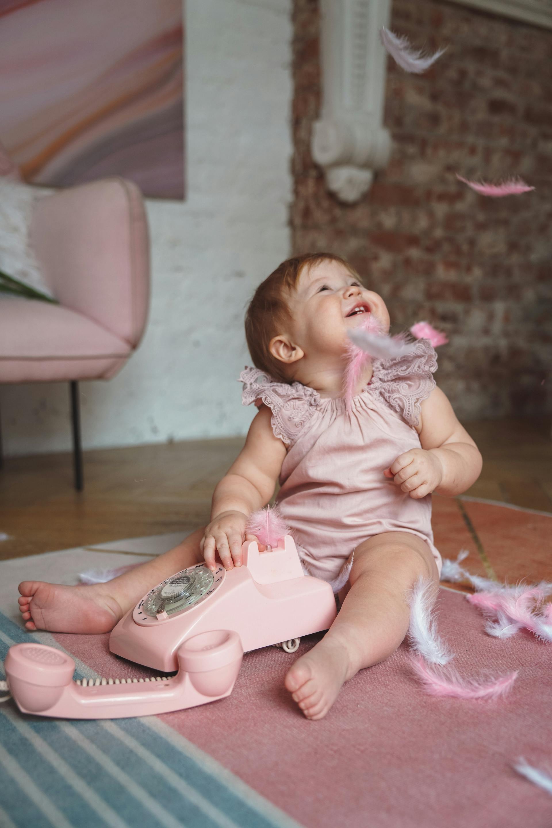 A baby girl dressed in pink clothing playing with feathers and a pink telephone