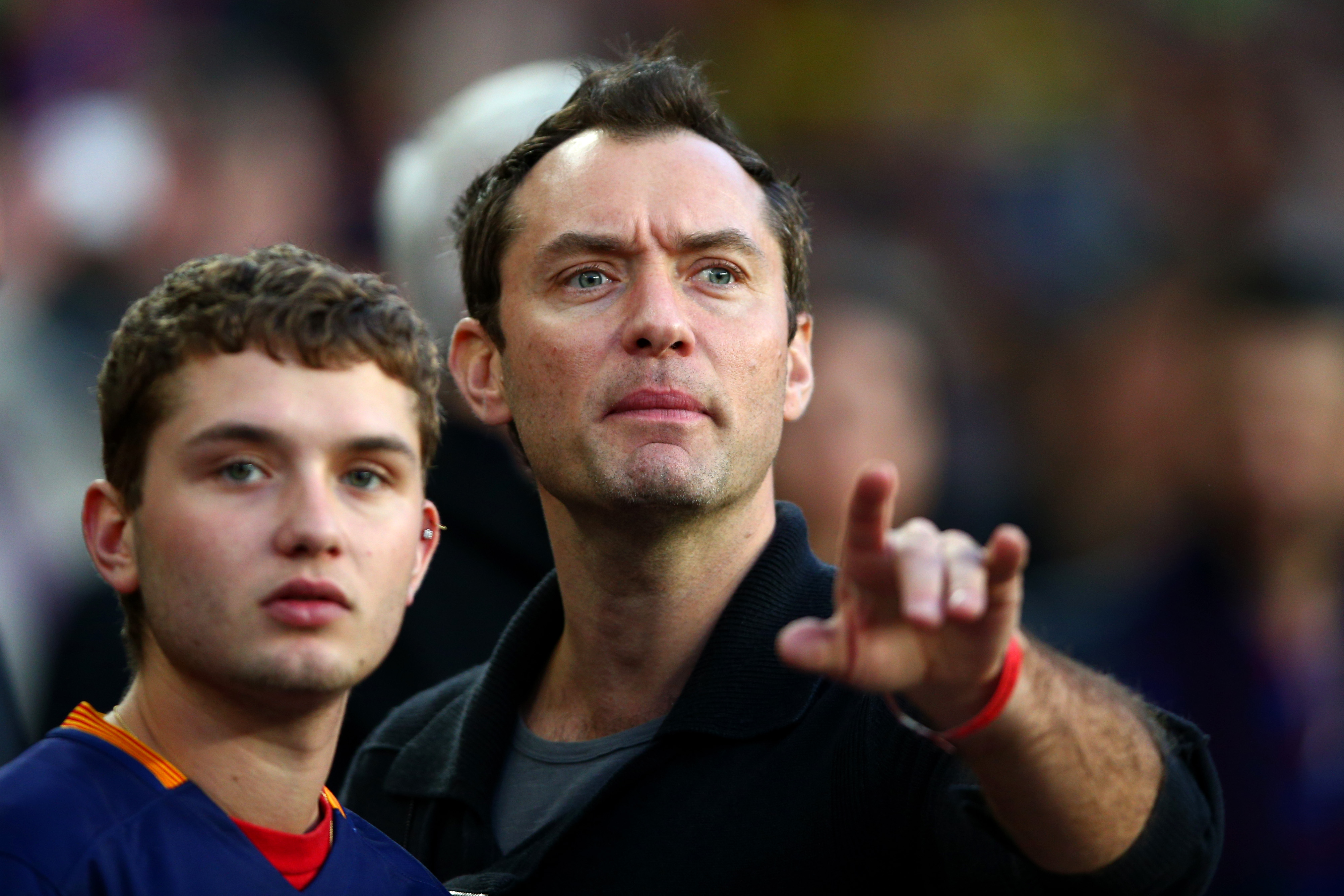 Jude Law and a teenage Rafferty in the stands at the Barcelona vs Real Madrid match at Camp Nou, April 2016 — Jude pointing ahead while Raff watches quietly beside him, the matching bone structure visible in profile.