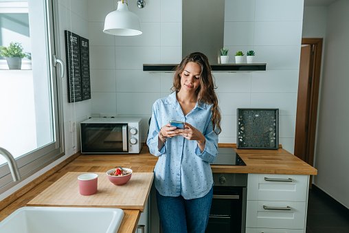 Photo of a young woman wearing pyjama in kitchen at home using cell phone | Photo: Getty Images