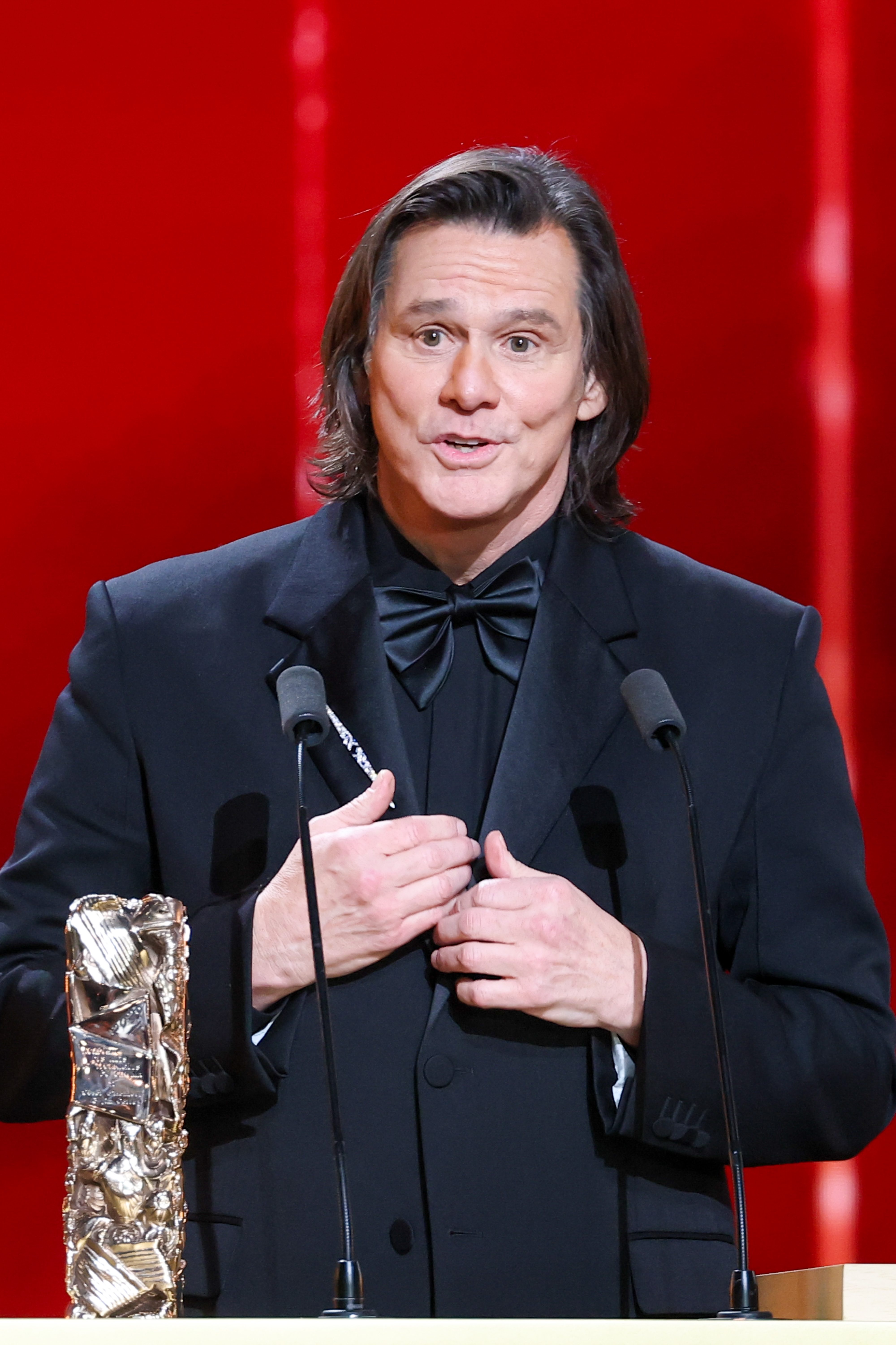 Dressed in a sharp black tuxedo, Jim Carrey stands at the podium with the Honorary César Award beside him. He gestures mid-speech between twin microphones against a bold red stage backdrop. The actor accepts the honor during the 51st César Film Awards.