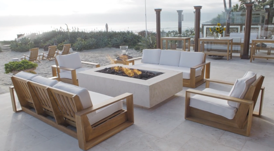A large travertine fire pit with flickering flames centers an inviting outdoor lounge area equipped with teak sofas and plush white cushions. Beyond the tiled patio, a sandy dune planted with succulents leads down toward the ocean shore under a hazy coastal sky. | Source: YouTube/Architectural Digest