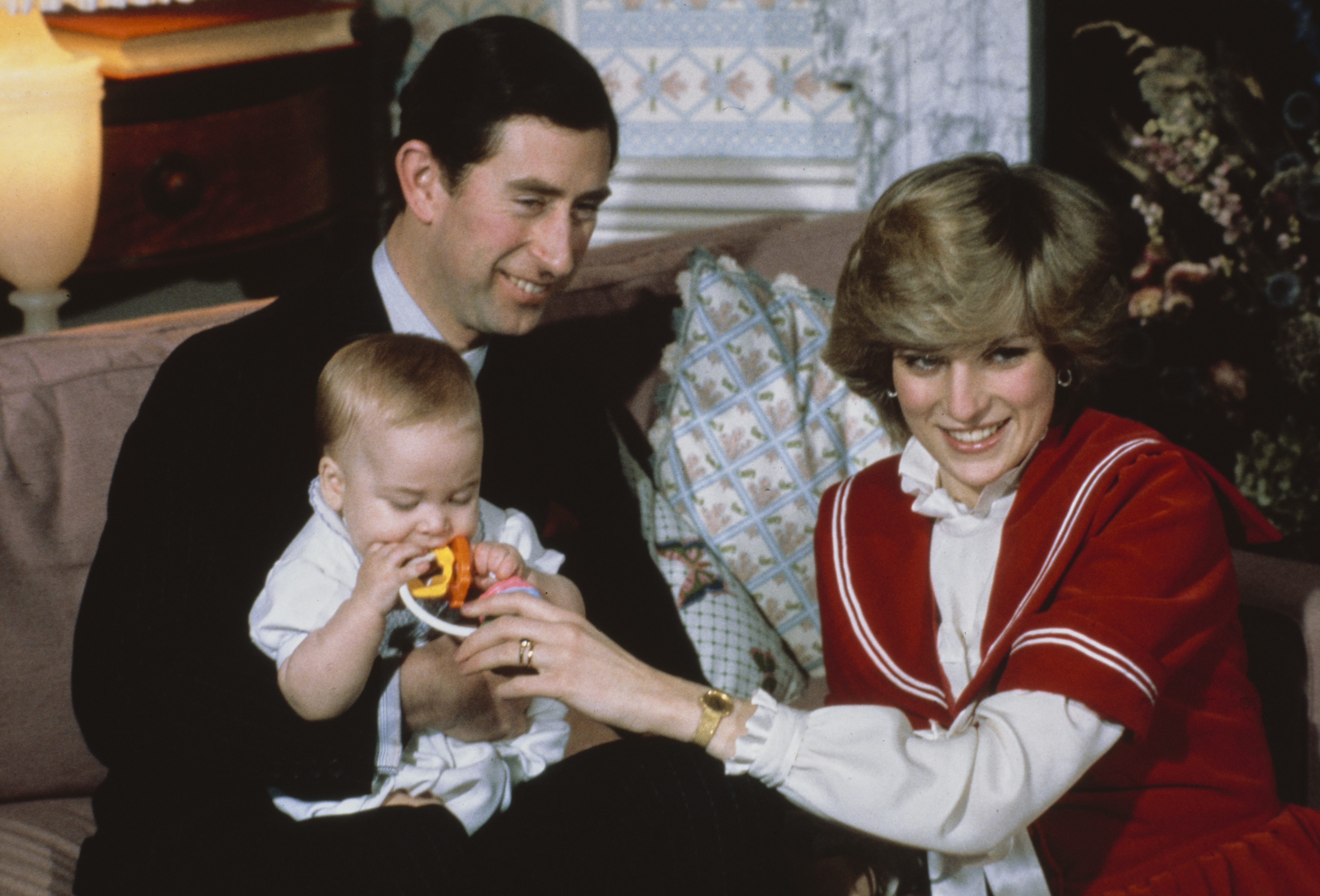 Captured at Kensington Palace on December 22, 1982, this tender family portrait shows King Charles III, then the Prince of Wales, cradling a young Prince William as Diana, former Princess of Wales, gently offers a teething ring. Dressed in a red-and-white sailor-style outfit, the Princess radiates youthful warmth and joy.