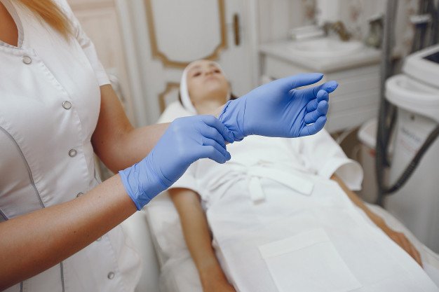 Mujer poniéndose guantes de látex mientras una paciente espera en la camilla. │Foto: Freepik