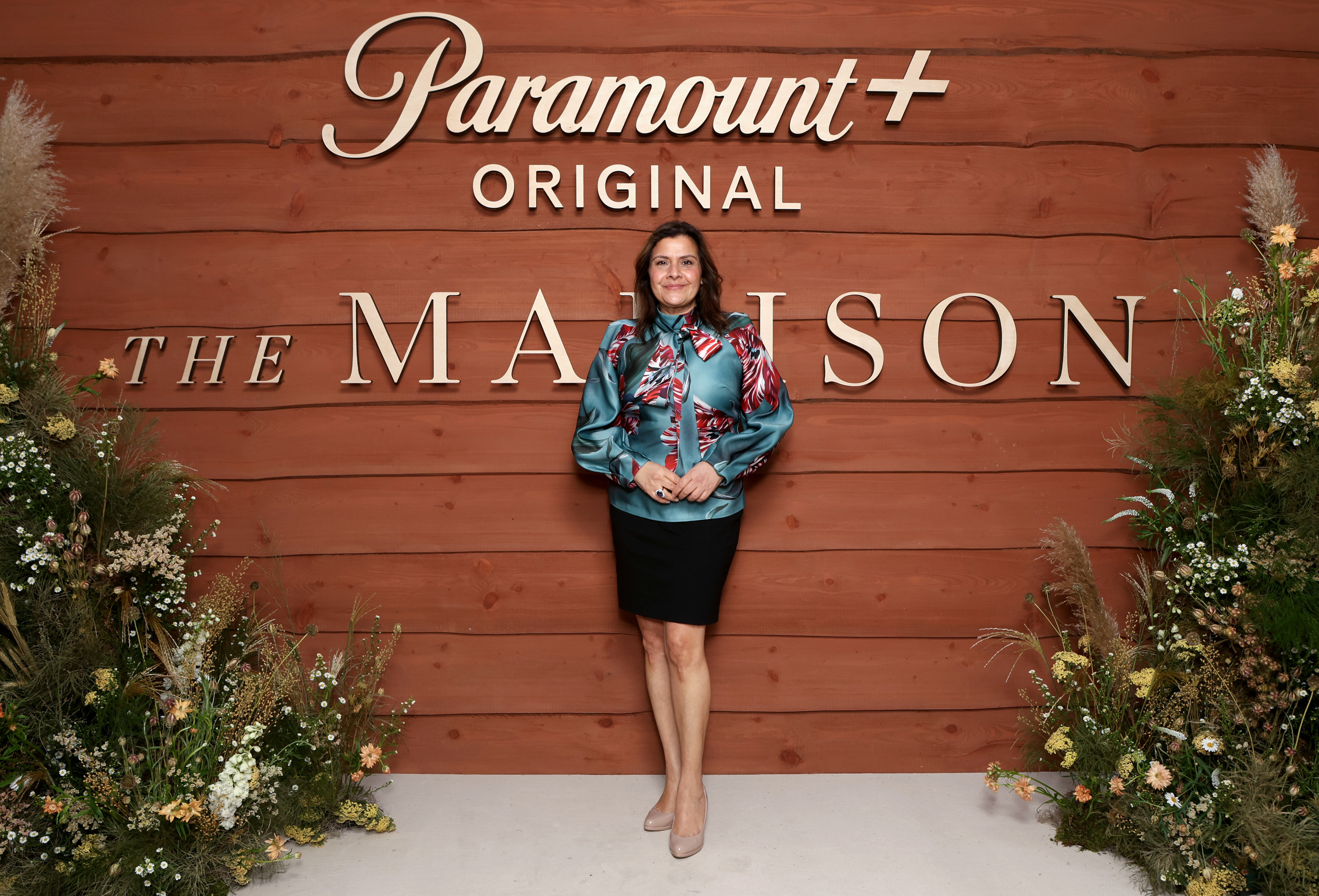 A visibly transformed Wadia poses in front of a warmly lit cedar wood backdrop flanked by lush arrangements of wildflowers and pampas grass at a Paramount+ lunch celebrating "The Madison" at 1 Hotel Mayfair in March 2026. She wears a teal satin blouse with a bold red and white botanical print and a pussy-bow neckline, tucked into a sleek black mini skirt, her legs long and lean in nude court heels — a silhouette strikingly different from photos taken less than a decade earlier.