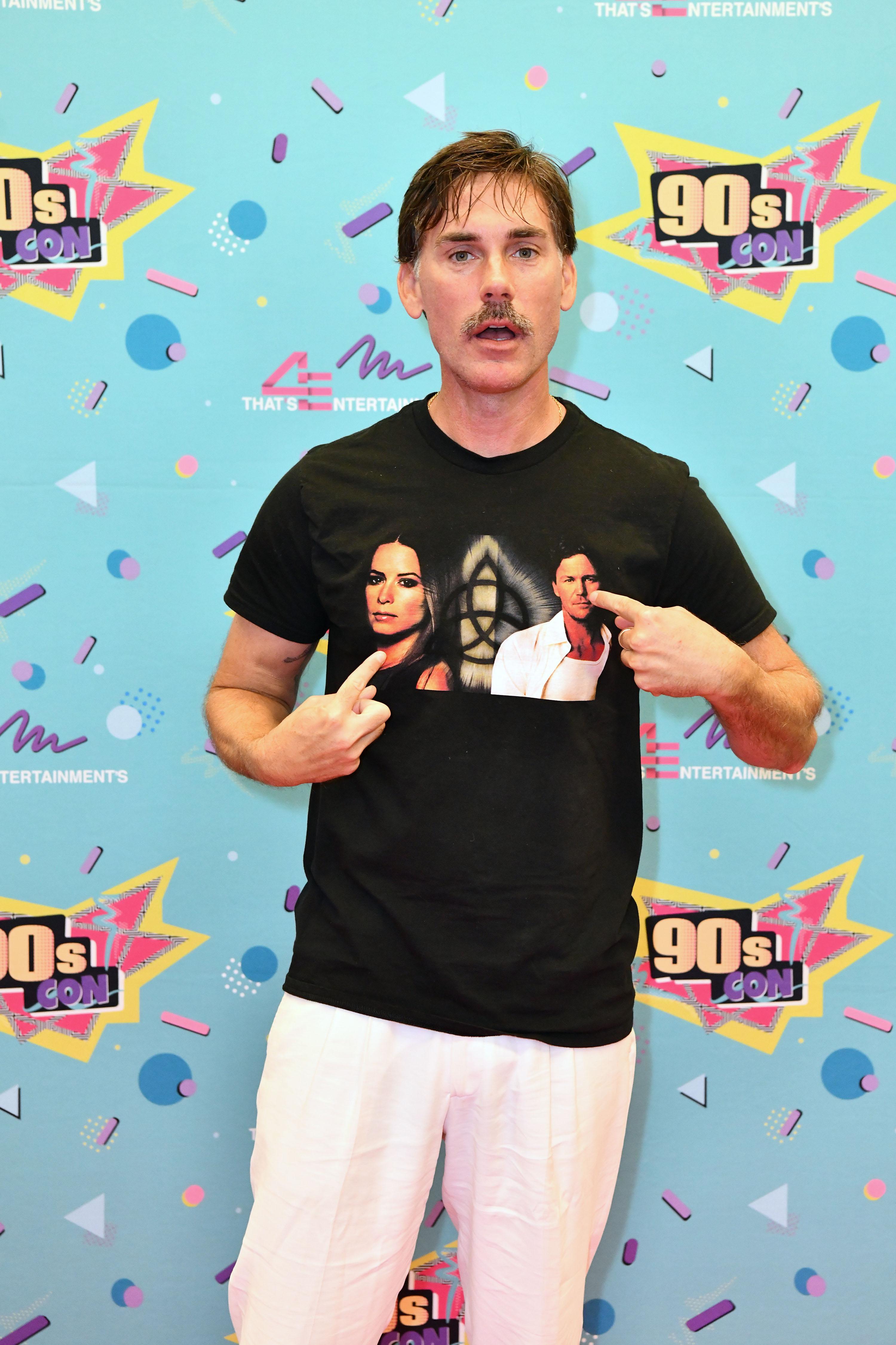 Drew Fuller makes a playful appearance at 90s Con in 2024, pointing to a custom T-shirt featuring his "Charmed" co-stars Holly Marie Combs and Brian Krause. Dressed in casual white pants and a black shirt, he poses with a humorous expression against a vibrant, retro-themed backdrop, embracing the nostalgic spirit of the event.
