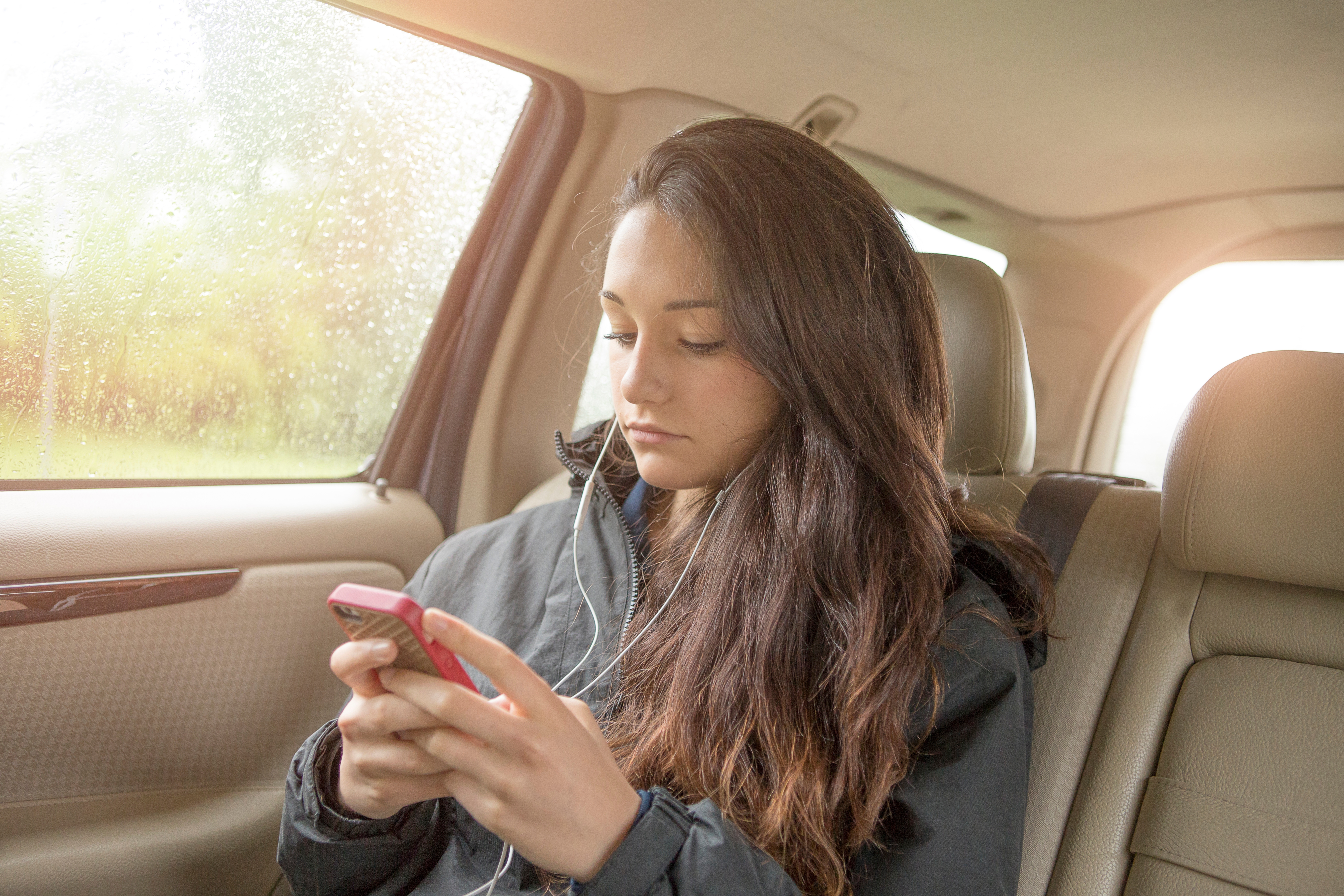 Sullen teenage girl selecting smartphone music in car back seat | Sources: Getty Images