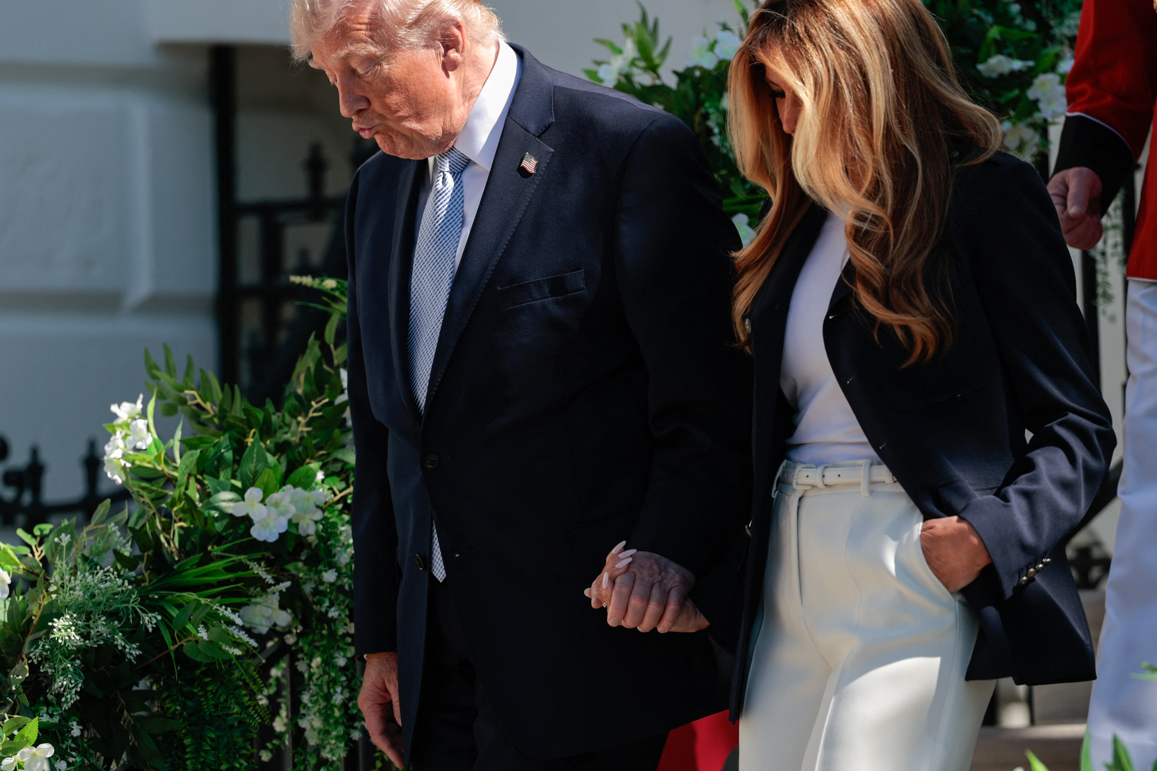 Donald and Melania Trump descend from the balcony hand in hand, surrounded by white floral displays, as they greet guests during the event on the South Lawn.