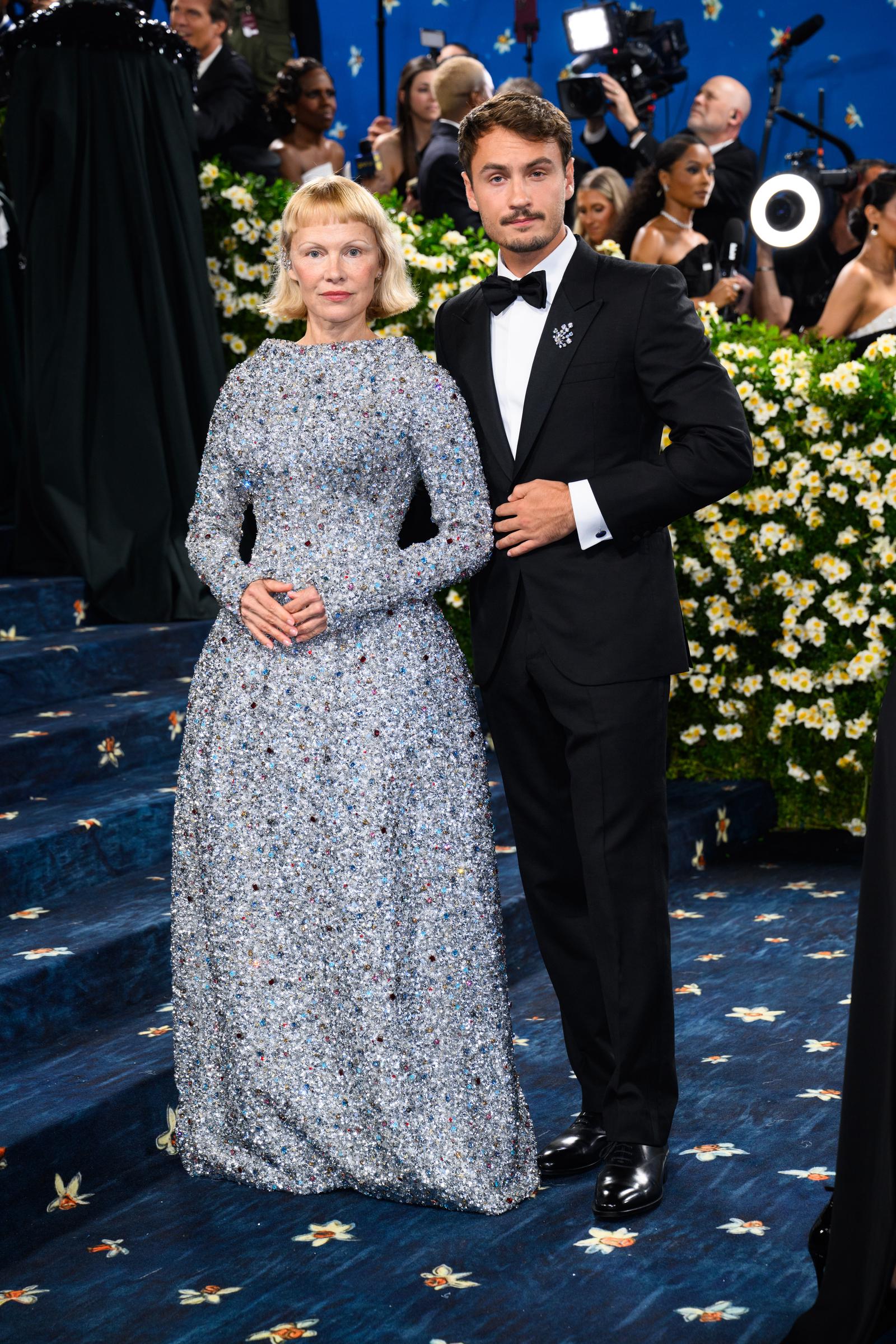 Surrounded by flashing cameras and a flurry of event coverage, Pamela Anderson and Brandon Thomas Lee pause near the iconic Met Gala staircase. Their composed presence stands out amid the lively backdrop of photographers and onlookers capturing the high-profile moment.