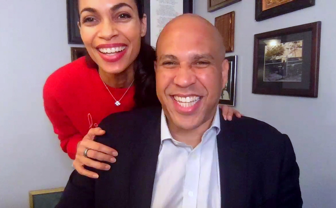Beaming with joy during a virtual appearance, Rosario Dawson stands behind Cory Booker as they laugh together on screen, capturing a playful and candid side of their relationship.