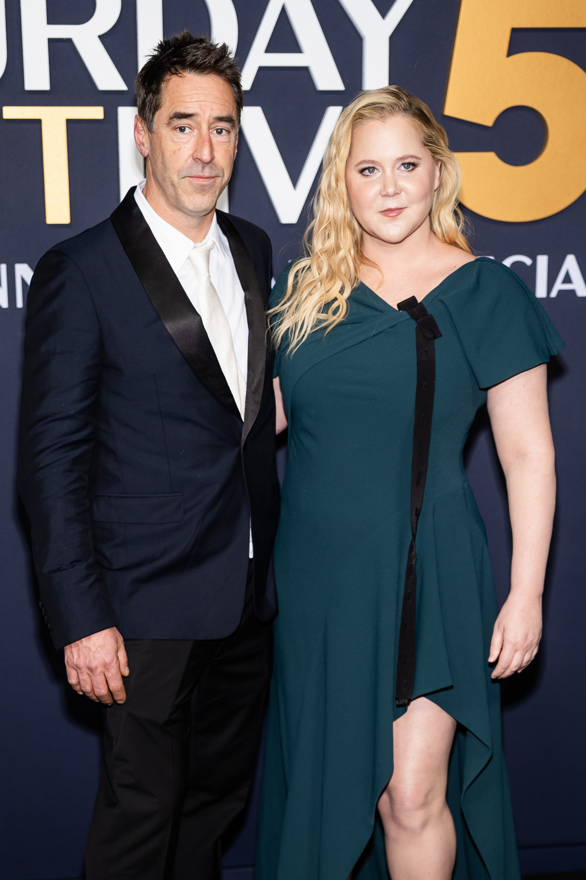 Amy Schumer posed alongside then-husband Chris Fischer at the "SNL50: The Anniversary Special" in New York City on February 16, 2025. Wearing a dark green asymmetrical dress with a thigh-high slit, Schumer appeared heavier through the arms and midsection, offering a clear visual contrast to her more recent, leaner frame shared online in 2026.