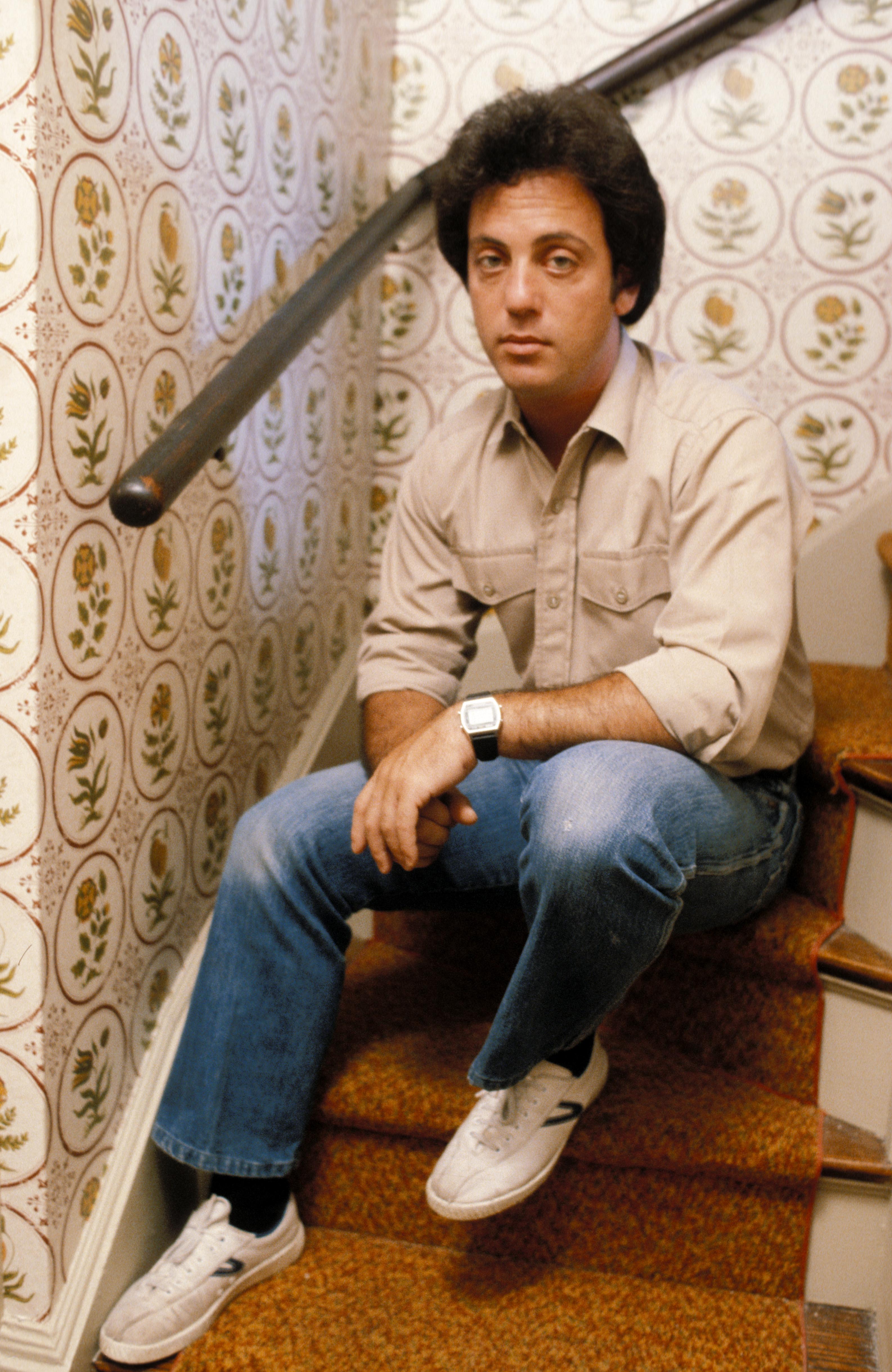 Rock & Roll Hall of Famer Billy Joel sitting on a staircase wearing a beige shirt paired with jeans and white sneakers. / Source: Getty Images