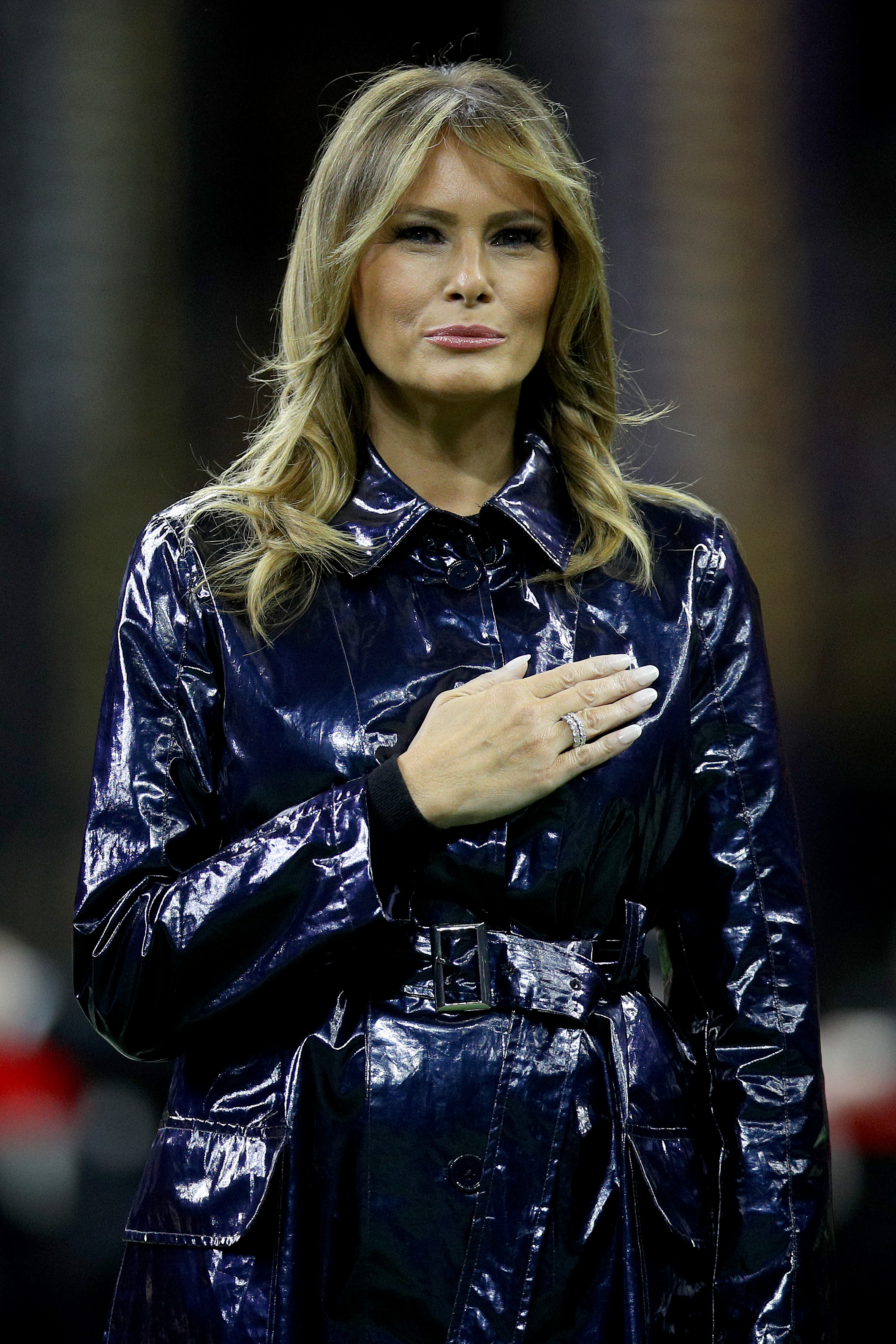 Melania Trump wore lighter blonde highlights and a glossy dark coat in this stadium appearance. The image shows a more dramatic look than earlier portraits in the gallery.
