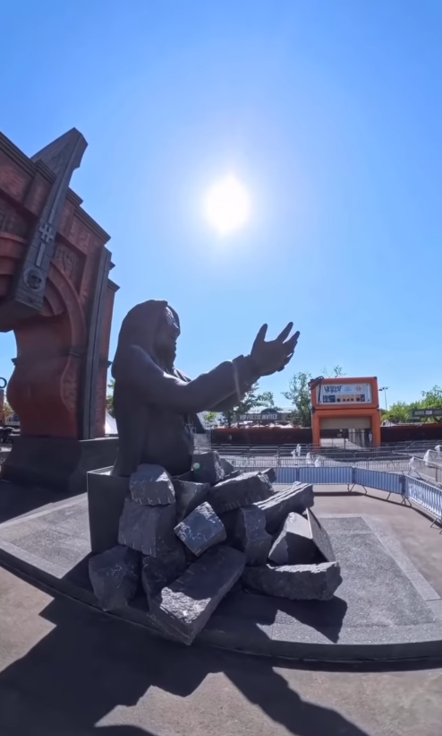 Seen in profile beneath the midday sun, Ozzy Osbourne's statue stretches its arms outward from a base of broken stone. Festival tents, barriers, and nearby buildings frame the tribute at Hellfest in Clisson, France. | Source: Instagram/sharonosbourne