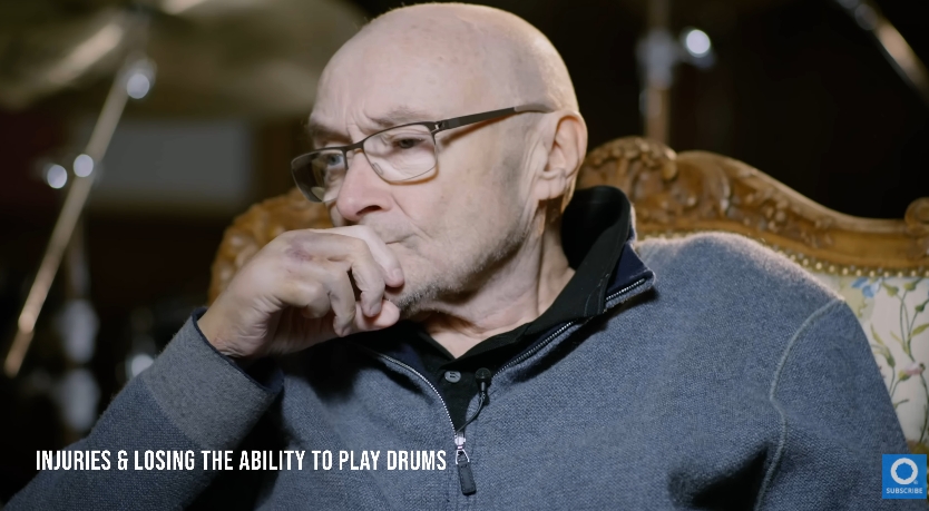 Phil Collins rests his hand against his face in a still moment of quiet thought. With visible signs of ageing and frailty, the image captures the toll taken by nearly two decades of health complications, but also the reflective strength of a man who continues to navigate life with grace after an extraordinary career. | Source: YouTube/Drumeo