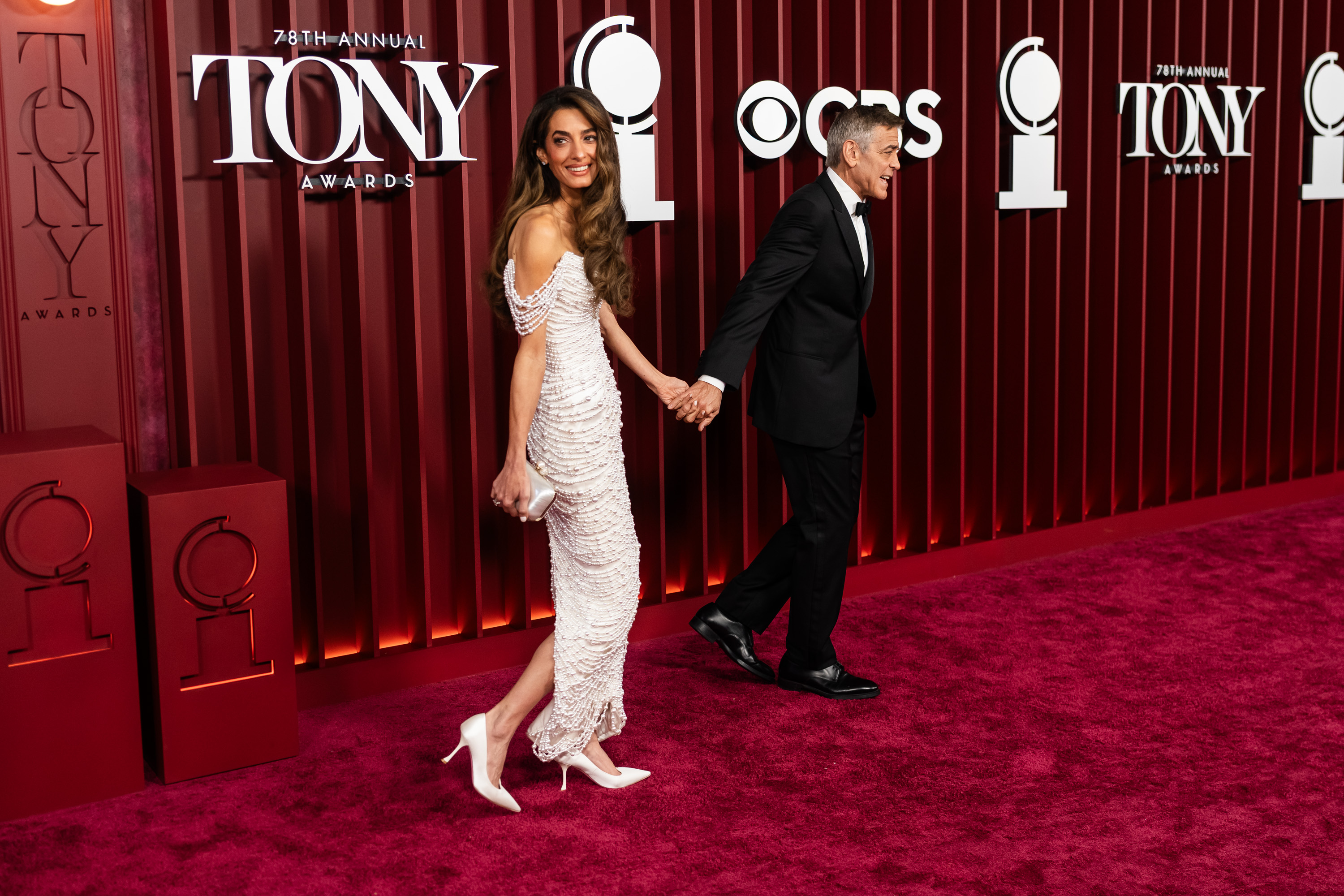George Clooney leads Amal Clooney down the red carpet at the Tony Awards, the pair exchanging expressions as cameras flash. The event's signature backdrop frames their poised arrival.