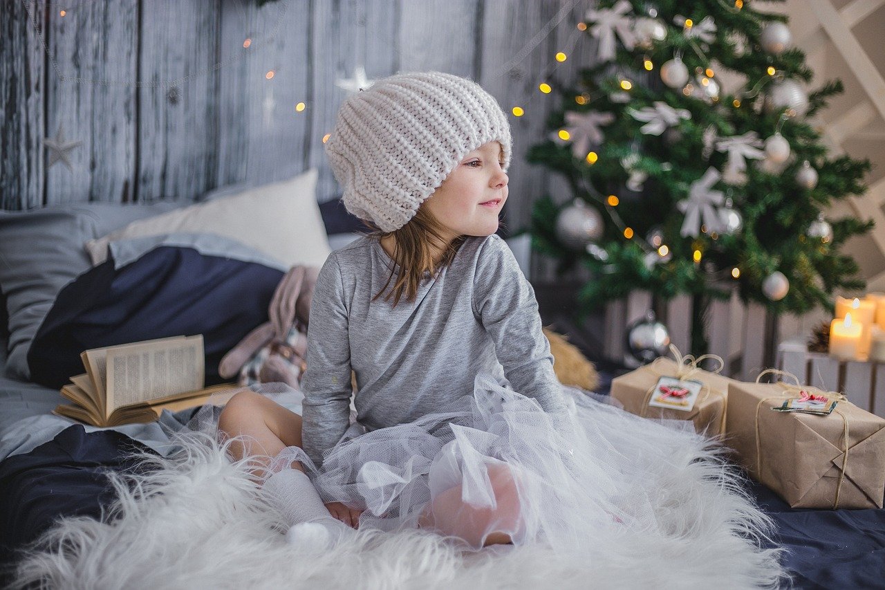 A little girl sitting on a bed and surrounded by Christmas presents, with a Christmas tree in the background. | Image: Pixabay.