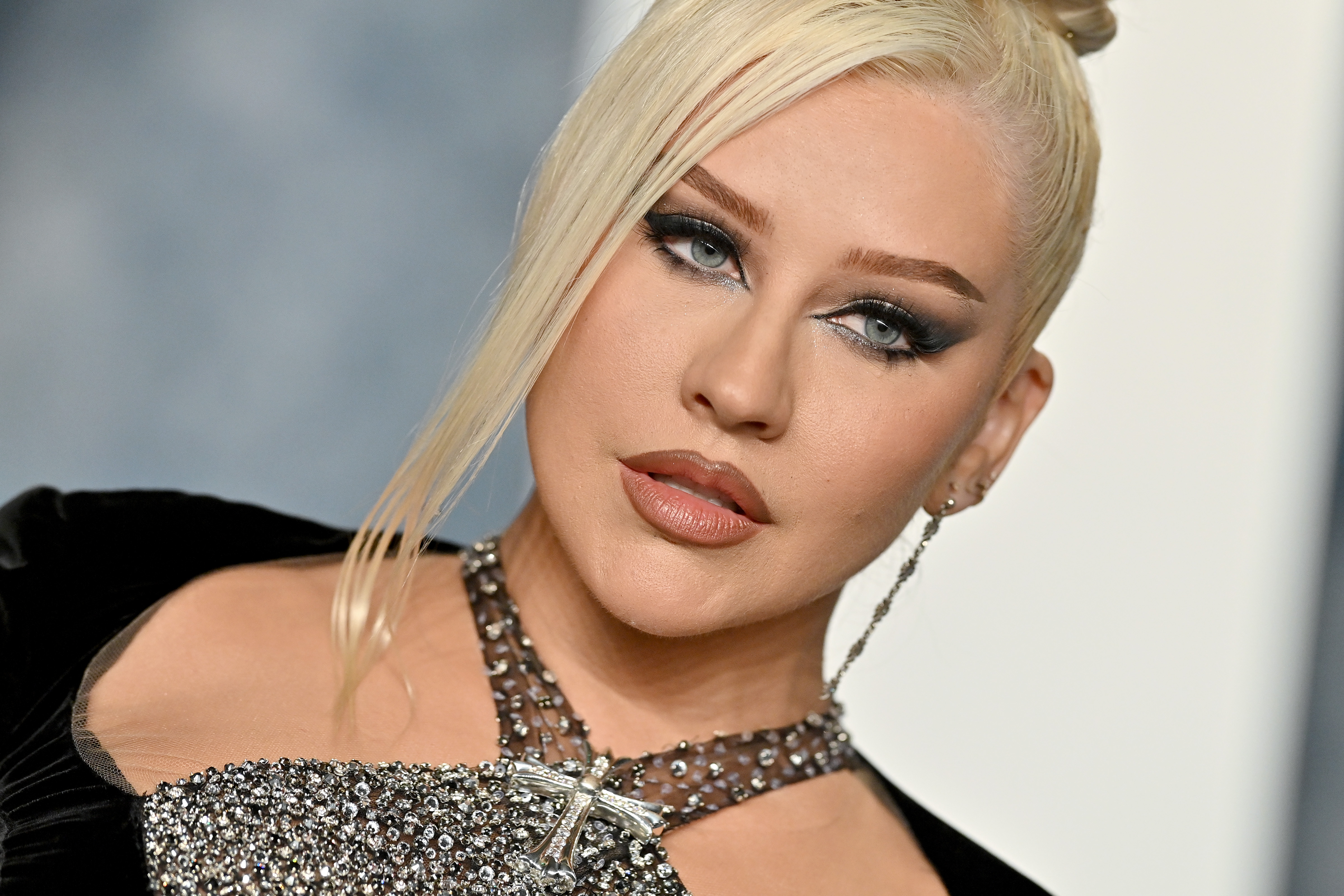 Christina Aguilera showcases a bold beauty look at the Vanity Fair Oscar Party in March 2023. Her makeup features smoky, winged eyeshadow, fluttery lashes, and a matte nude lip. She wears a dangling chain earring on one side and a cross-embellished crystal halter neckline, with her platinum blonde hair styled in a sleek topknot and a sharp face-framing strand.