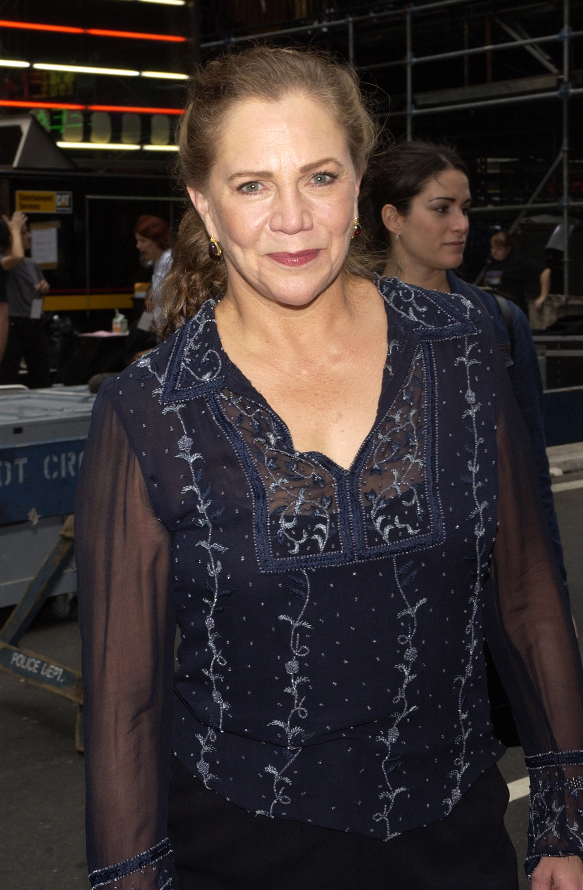 Kathleen Turner is pictured at the "Broadway on Broadway" event in Times Square, New York City. She wears a navy sheer embroidered blouse with delicate detailing, paired with dark trousers, radiating a polished yet approachable stage presence.