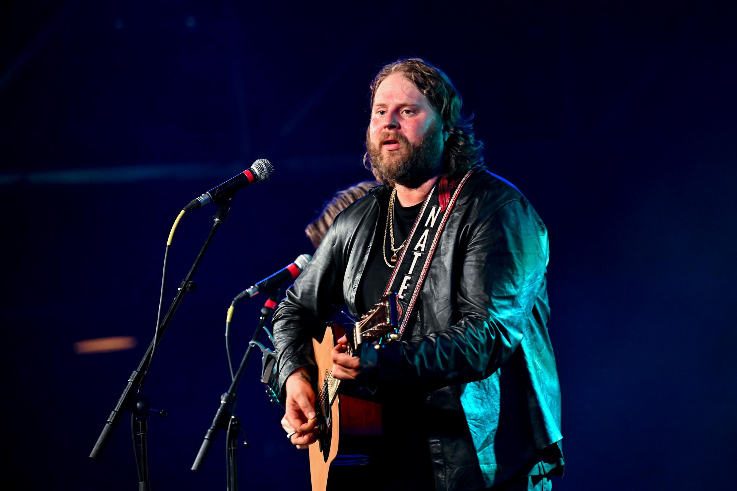 Nate Smith delivers an intense performance under blue stage lights, singing into the microphone while strumming his acoustic guitar in a black leather jacket.