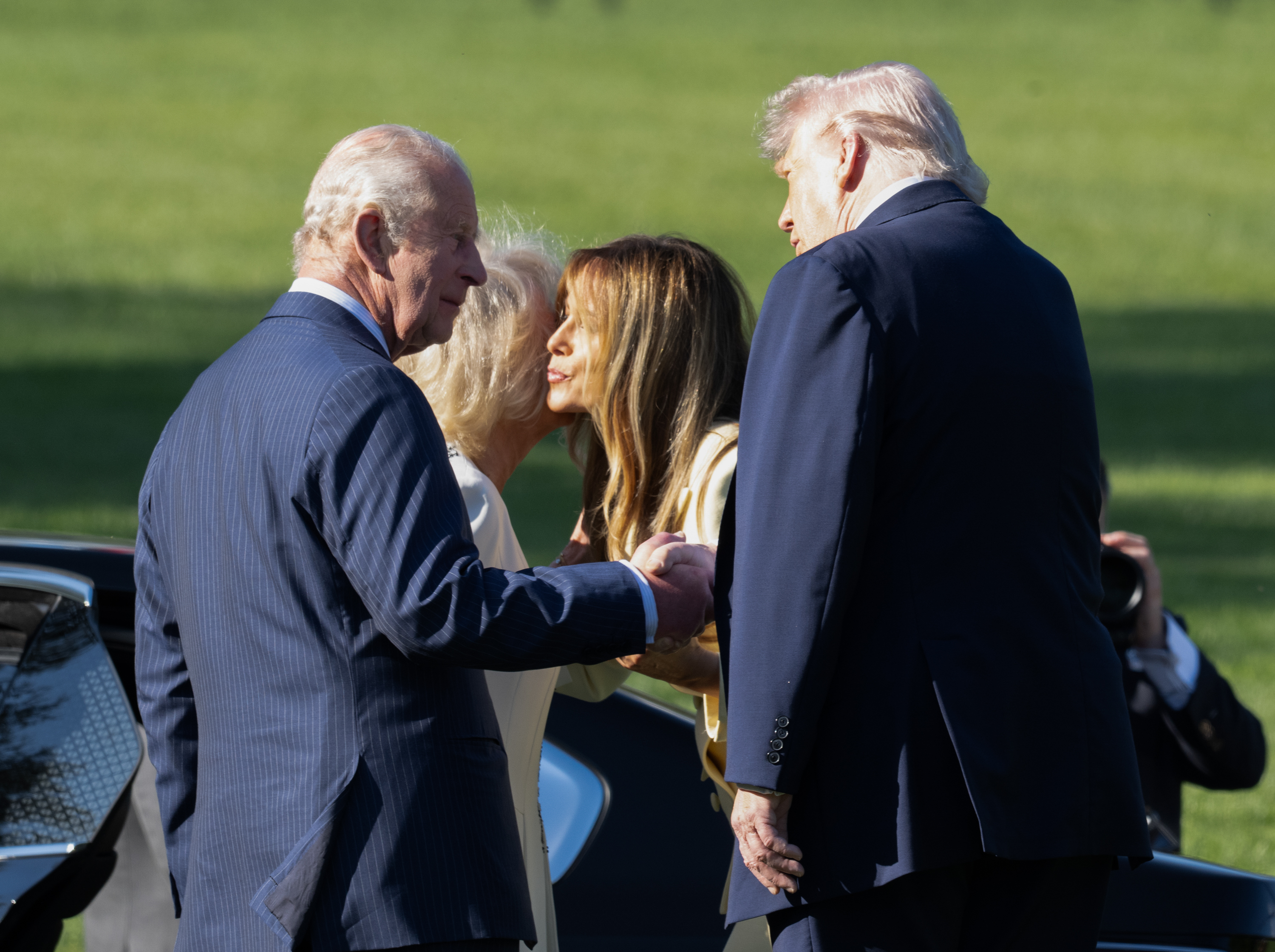 King Charles III and U.S. President Donald Trump appeared to exchange a firm handshake as First Lady Melania Trump greeted Queen Camilla with a cheek-to-cheek kiss beside the waiting vehicle on the White House lawn on 27 April 2026 in Washington, D.C. The overlapping greetings captured a moment of layered diplomacy, as both couples navigated formal protocol and personal courtesies at the start of the state visit.