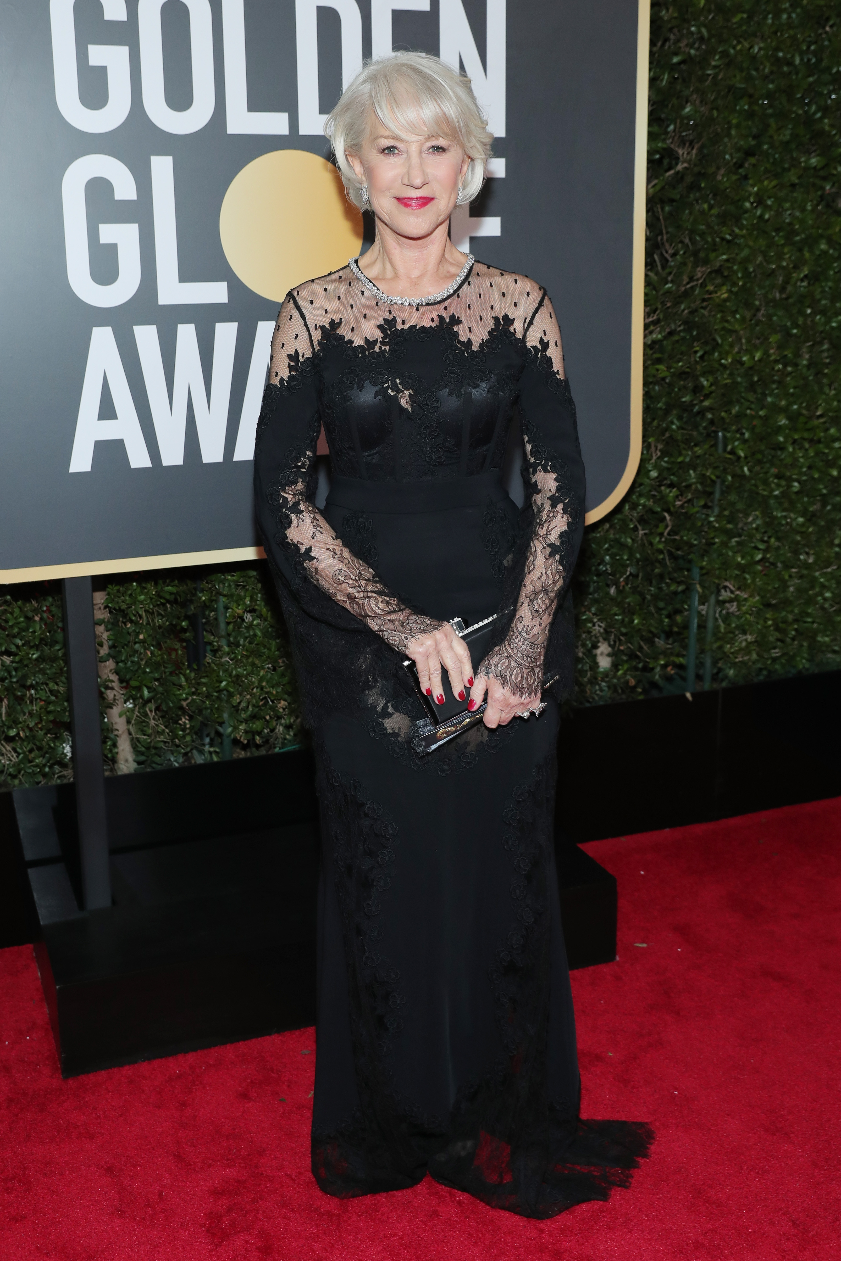 Helen Mirren captivates on the Golden Globe Awards red carpet in a floor-length black gown with sheer lace sleeves and an illusion neckline, accessorizing the elegant look with a silver necklace, a matching clutch, and a bold red lip to complete her timeless ensemble.