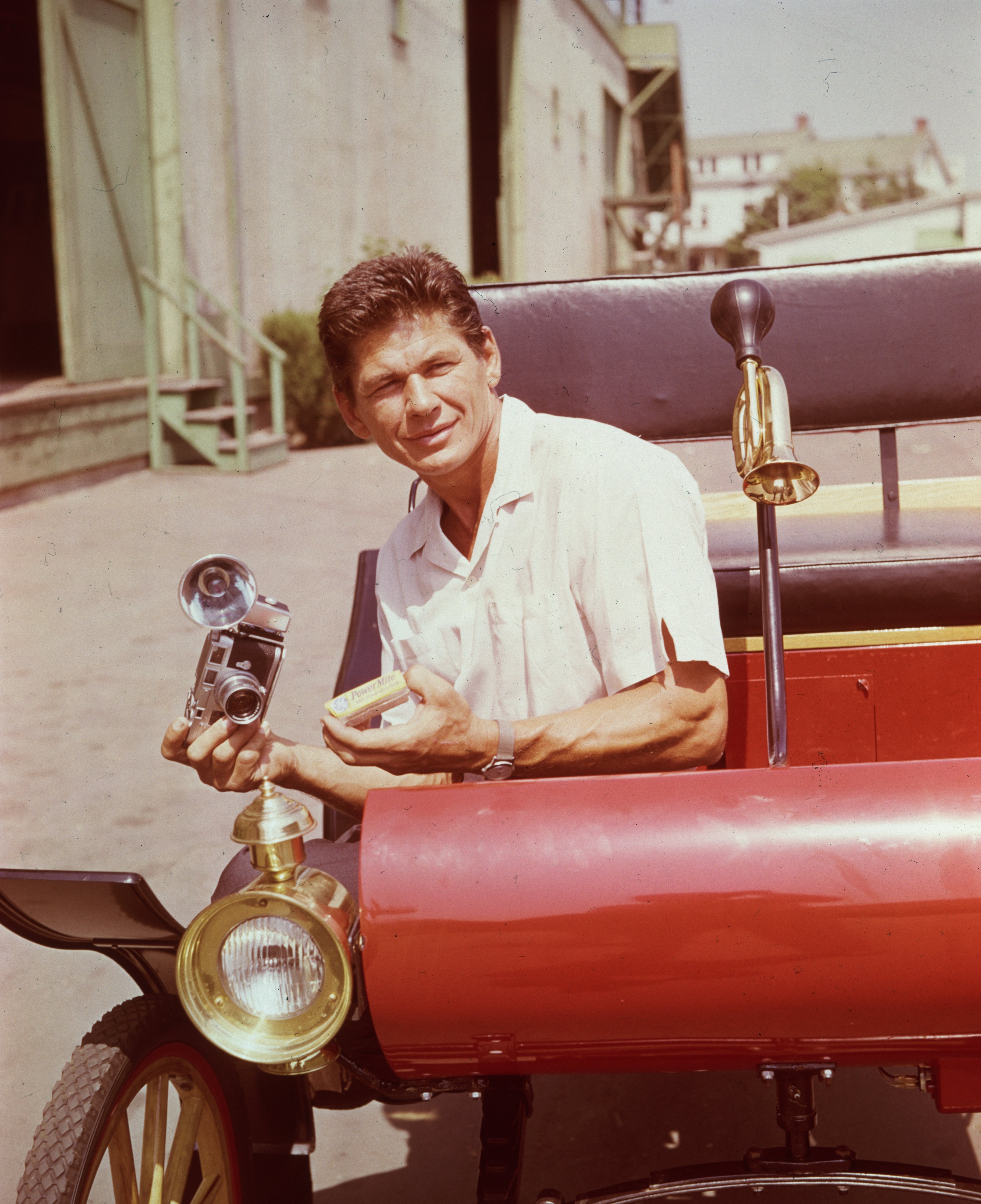 Charles Bronson (1921 - 2003) sits in the front of a 1920s automobile, holding a Leica flash-camera, on a studio backlot, California. | Source: Getty Images