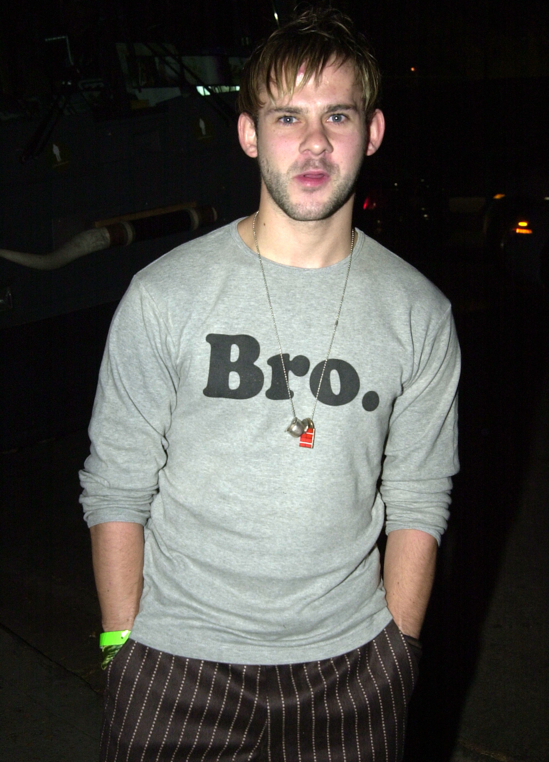 Dominic Monaghan attends the Rock Da House concert during Fashion Week LA in 2003 at Quixote Studios in West Hollywood, California, wearing a grey long-sleeve shirt with bold lettering, striped trousers, and a casual necklace, his slightly open-mouthed expression giving him a relaxed, edgy look.