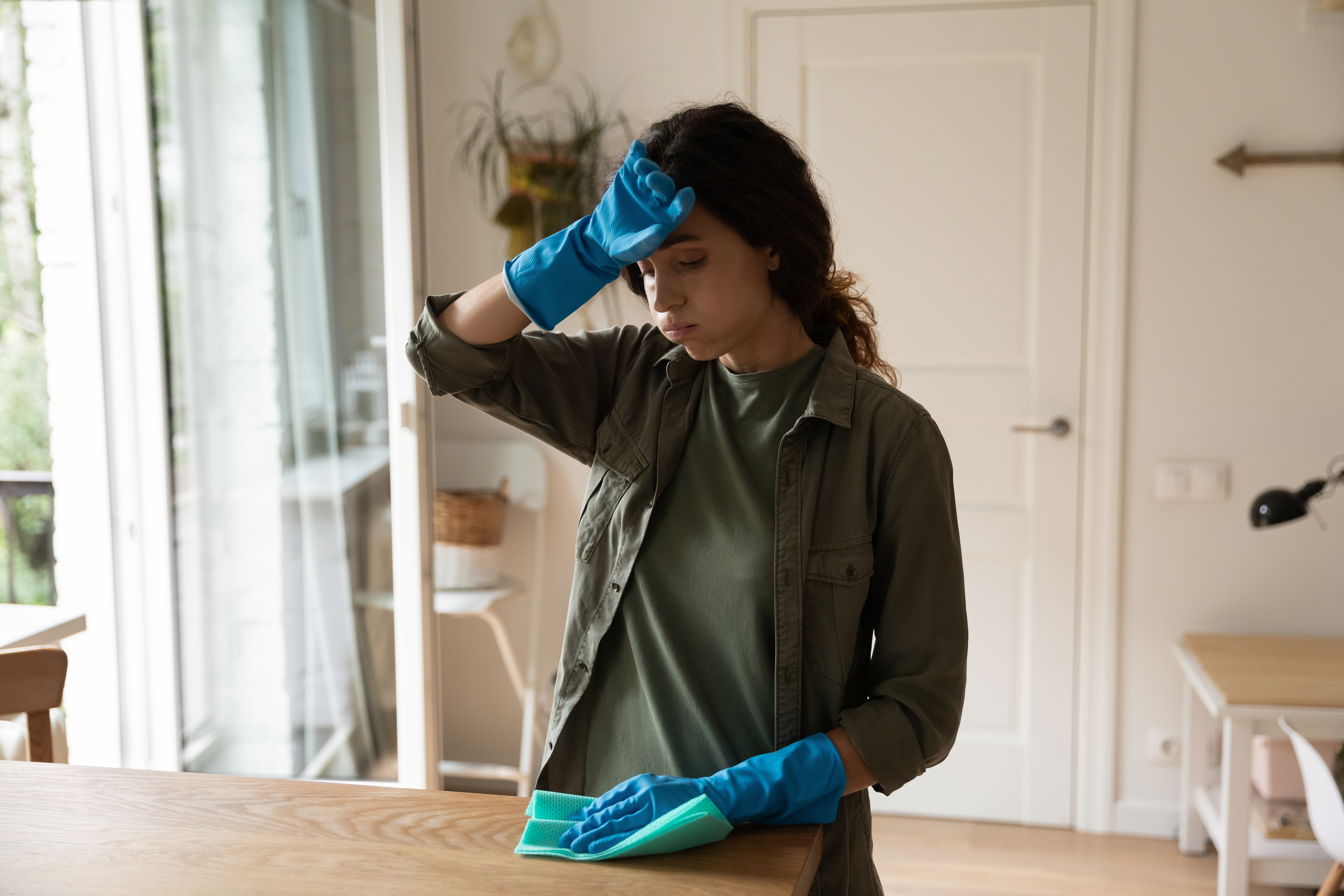 Exhausted millennial housewife in blue latex gloves wipe sweat from brow. | Source: Shutterstock