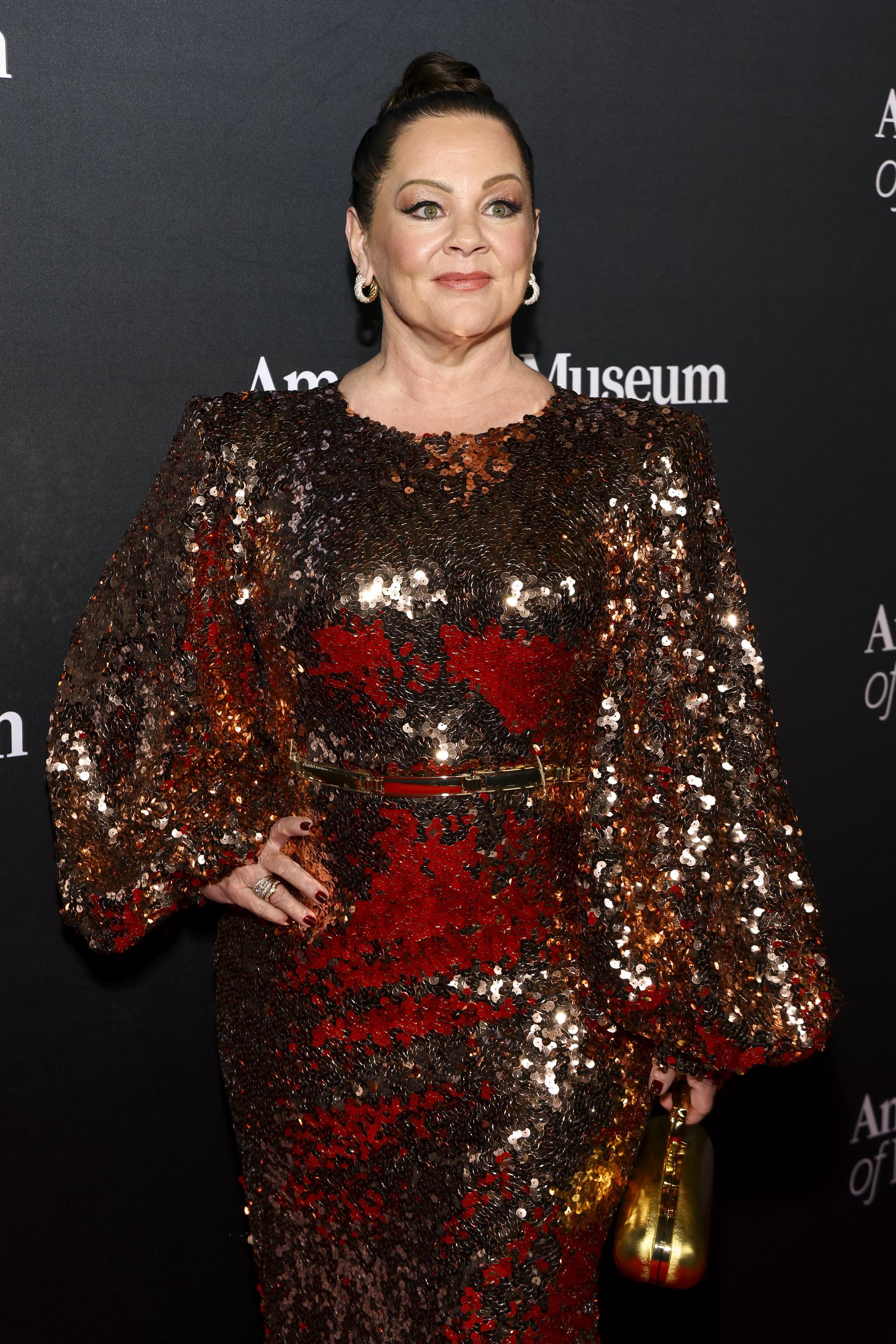 Melisa McCarthy stands before the event backdrop in a shimmering mosaic of bronze and deep red sequins, the gown's dramatic sleeves catching every glint of light. With a sleek updo and a metallic clutch in hand, she delivers a bold, statuesque elegance fit for a museum gala.