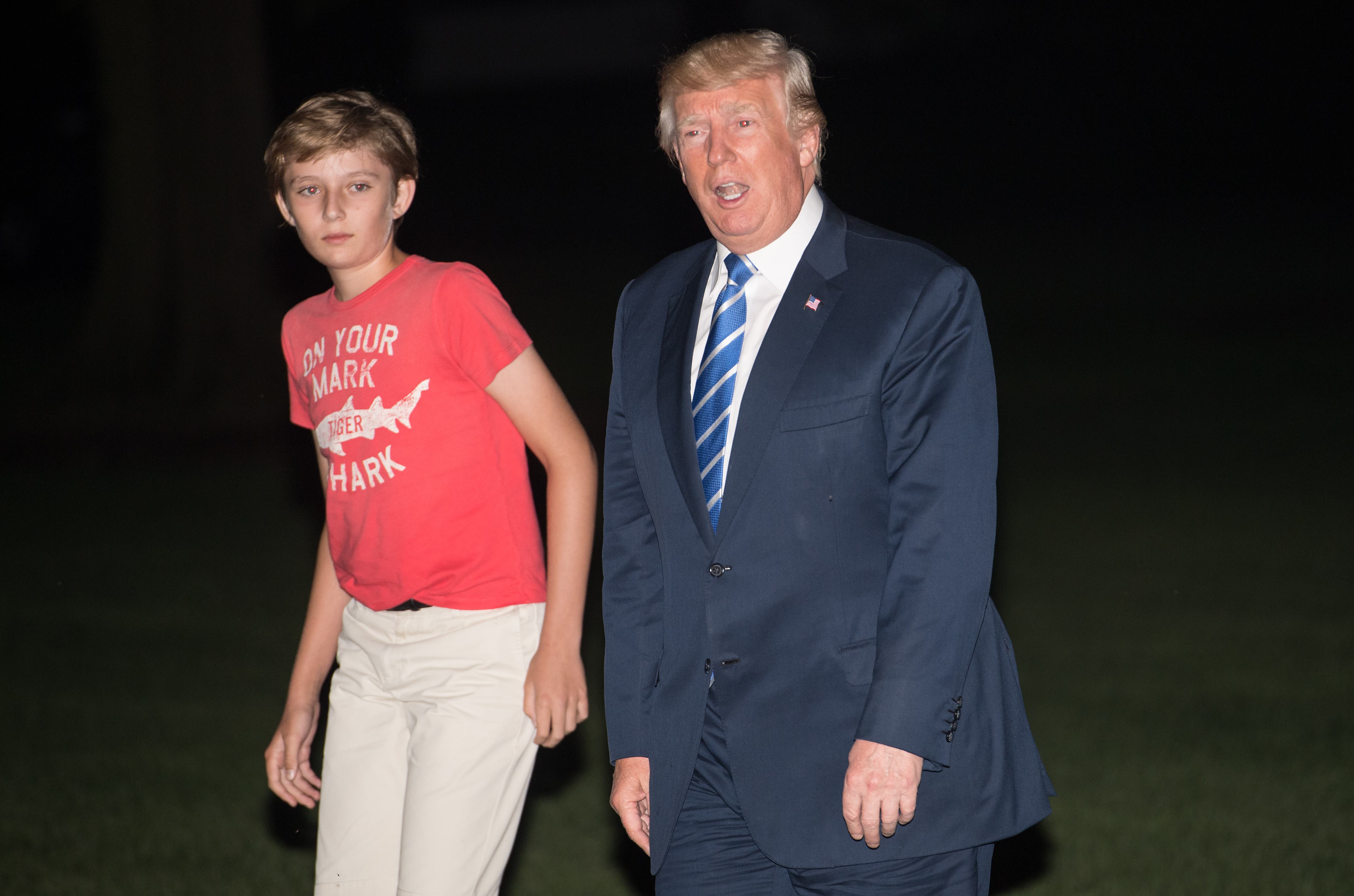 Donald Trump walks alongside his son Barron as they return to the White House in Washington, D.C. Barron keeps it casual in a red graphic T-shirt and khaki pants beside his suited father. The pair appear side by side during the nighttime arrival on the White House grounds.