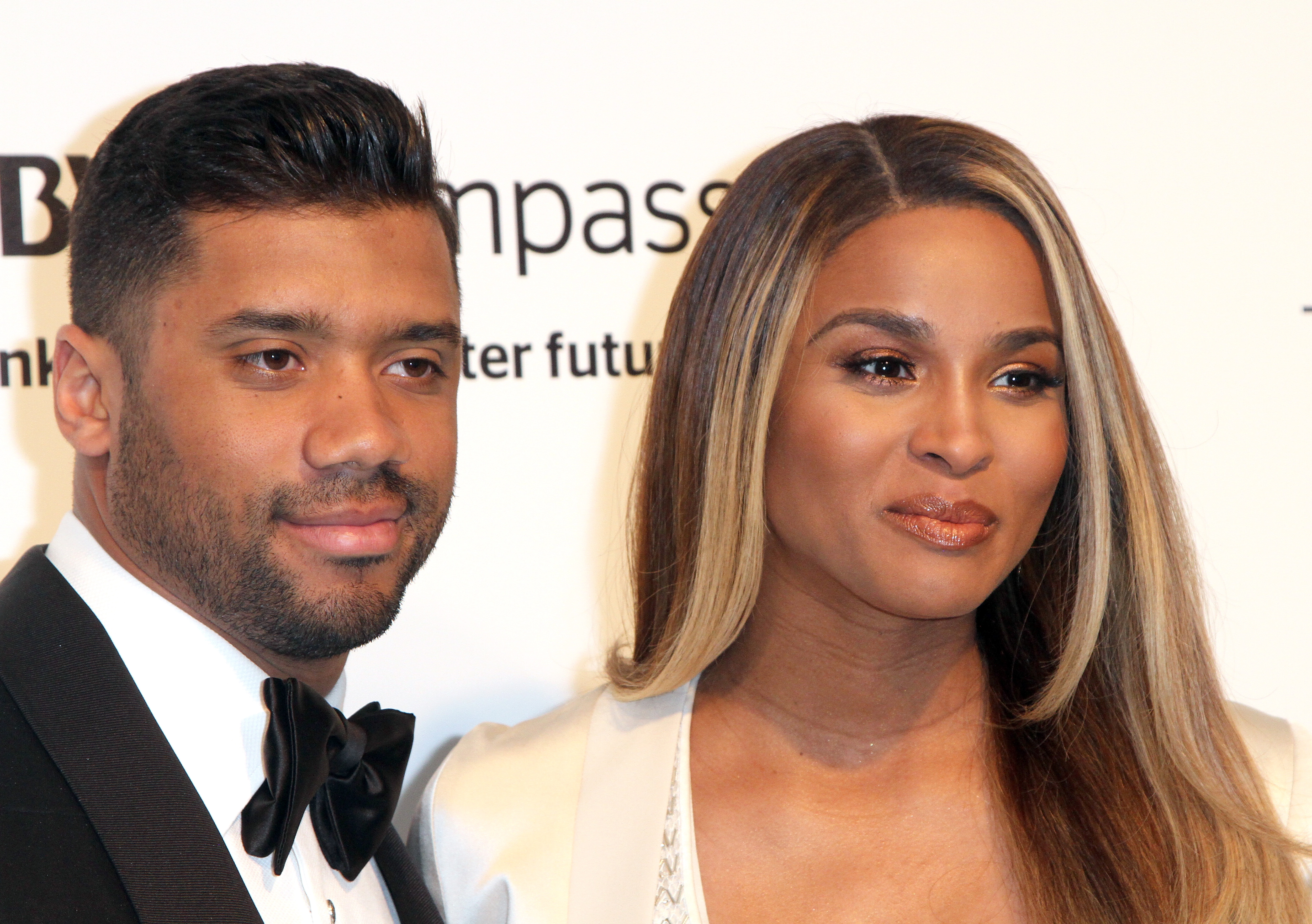 Ciara wears an elegant one-shoulder black gown as she stands beside Russell Wilson, who is dressed in a sharp, tailored tuxedo. Both face forward with calm, composed expressions, exuding a refined and understated red carpet presence.