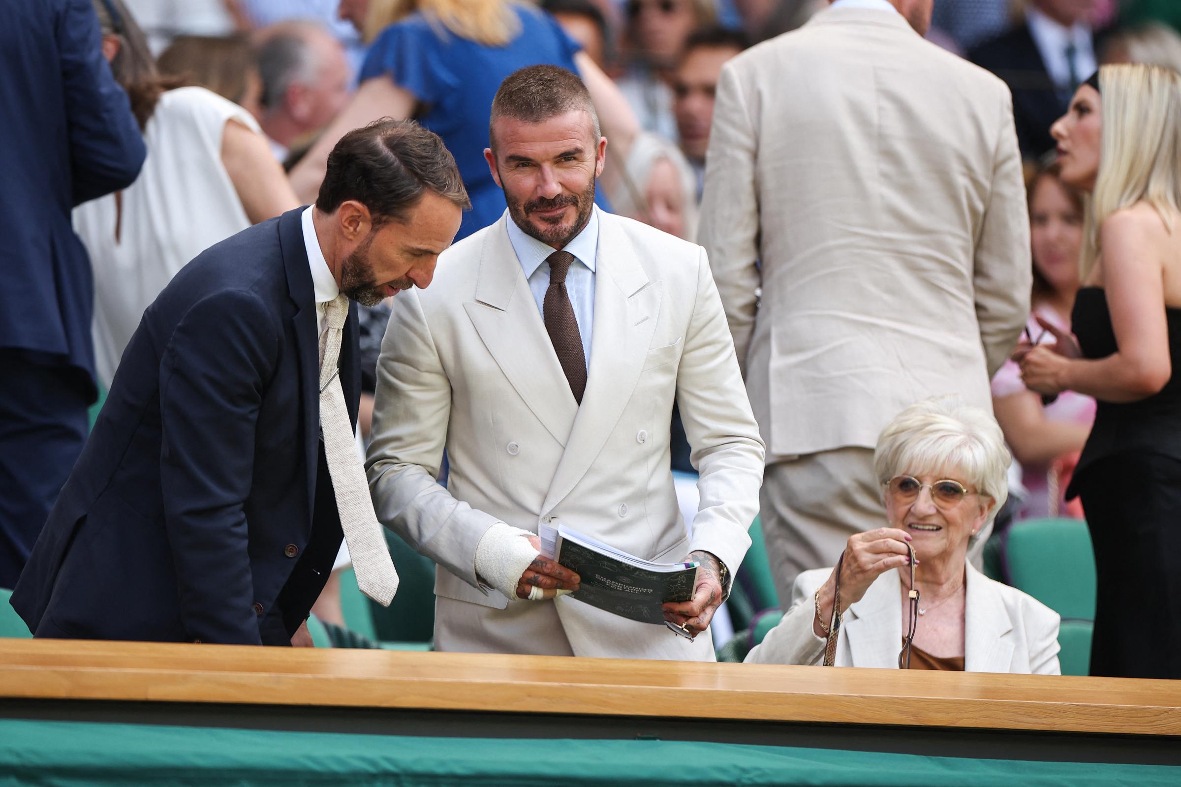 David Beckham stands beside Gareth Southgate, holding what appears to be a match programme as they converse before taking their seats. Meanwhile, Sandra Georgina West, seated nearby, smiles warmly while adjusting her glasses