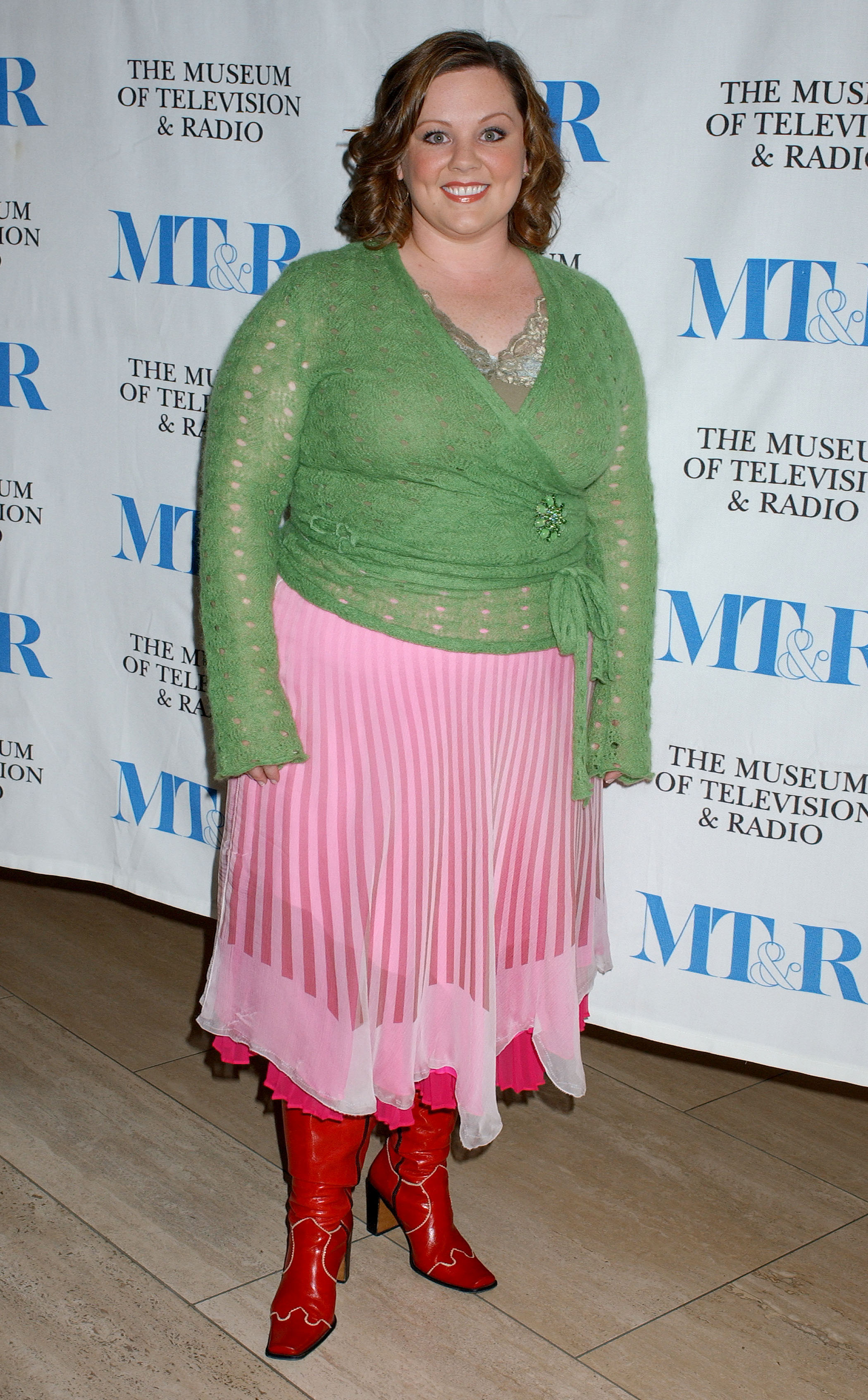 Melissa McCarthy appears smiling and vibrant, wearing a green knit top paired with a flowing pink skirt that adds a playful burst of color. Bold red boots complete the look, giving her outfit a lively, standout flair at the event.