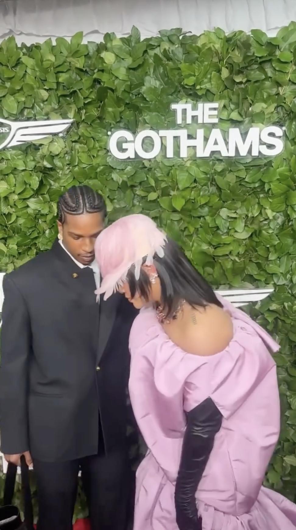 Rihanna adjusts the bottom of her dress, possibly to move more easily, while A$AP Rocky stands beside her. They focus on the moment in front of the Gotham Awards backdrop. | Source: Instagram/people