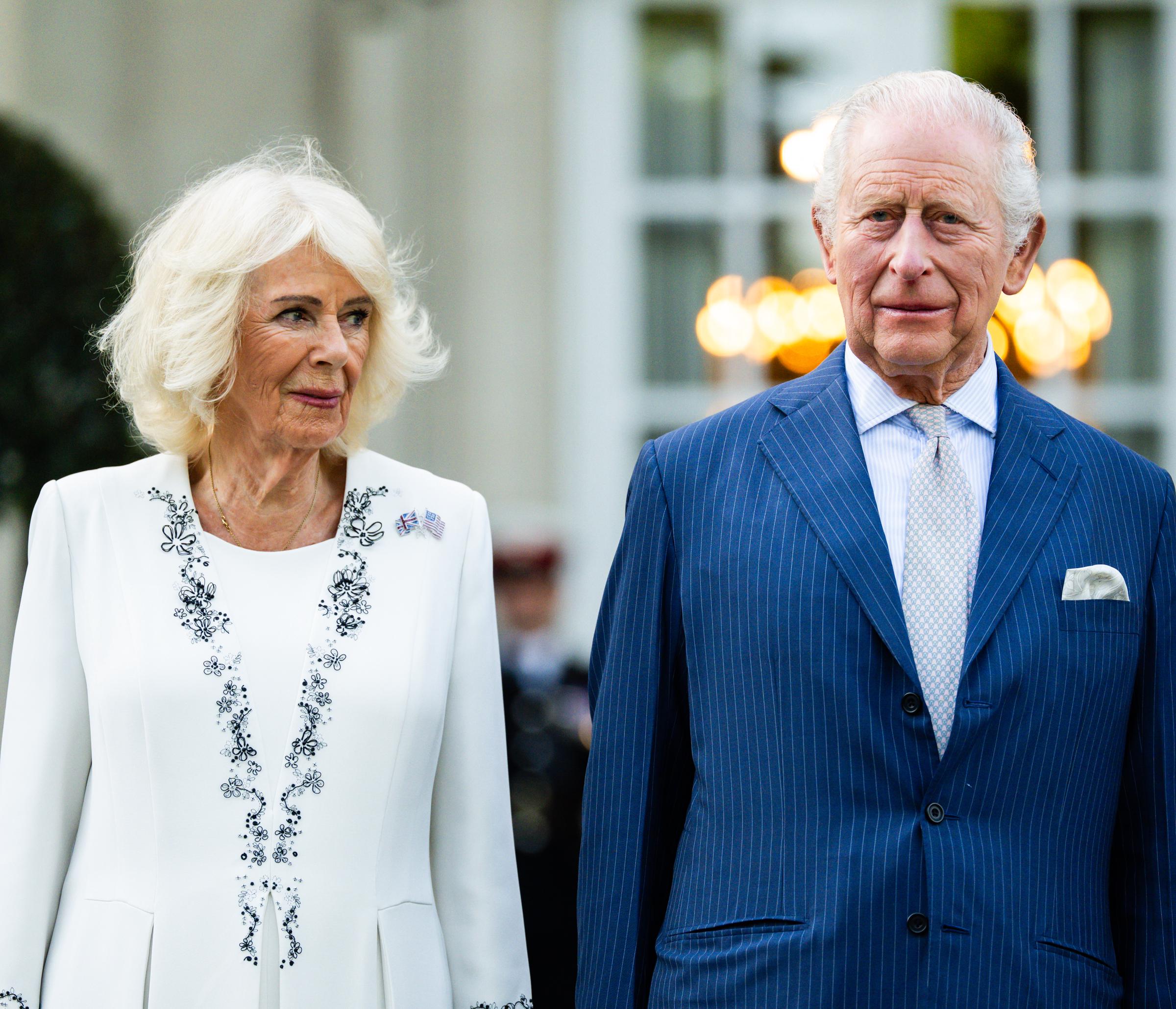 Queen Camilla and King Charles III were pictured standing side by side during a garden party on the White House grounds, with the Queen's embroidered white dress and the King's tailored navy suit offering a polished contrast against the softly lit backdrop. The couple appeared composed and attentive as they took in the occasion, part of a wider programme marking the beginning of their state visit celebrating the United States' 250th anniversary of independence.