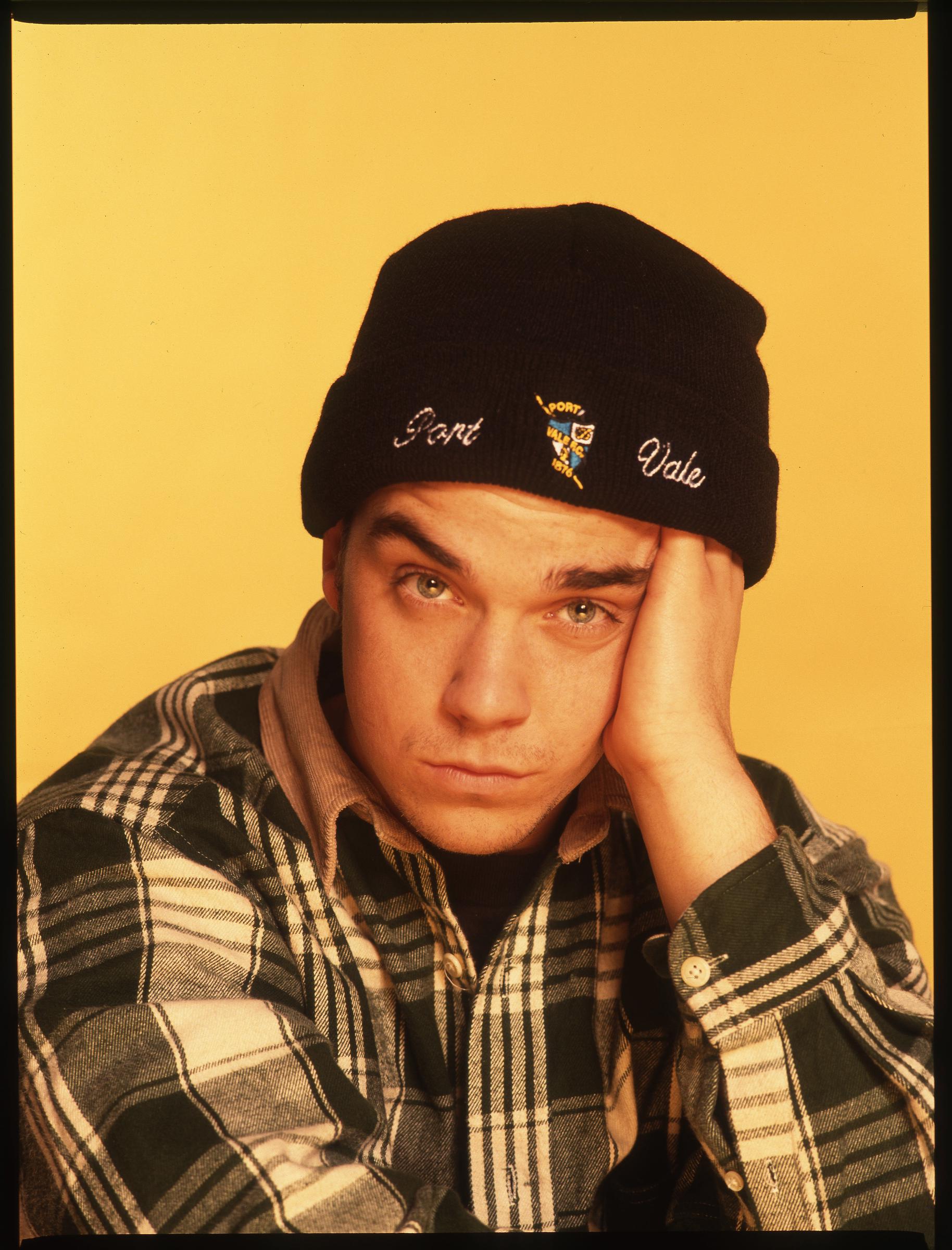 Robbie Williams is photographed in March 1993, resting his head in his hand while wearing a Port Vale beanie and green flannel shirt. The image captures a rare moment of stillness from the rising star, whose mix of attitude and vulnerability was already winning over fans.