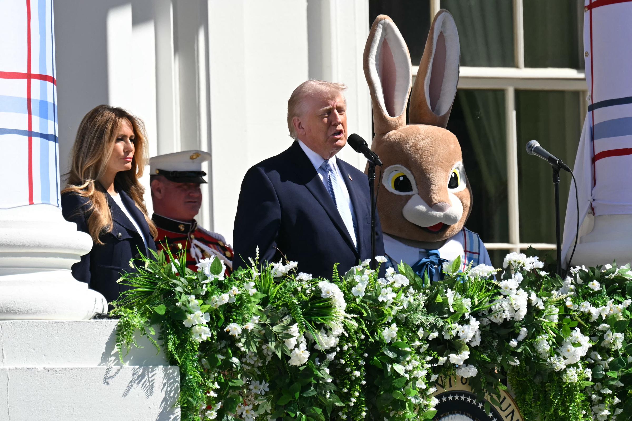 Donald Trump addresses the crowd from the White House balcony while Melania Trump and the Easter Bunny stand beside him, with the presidential seal partly visible below.