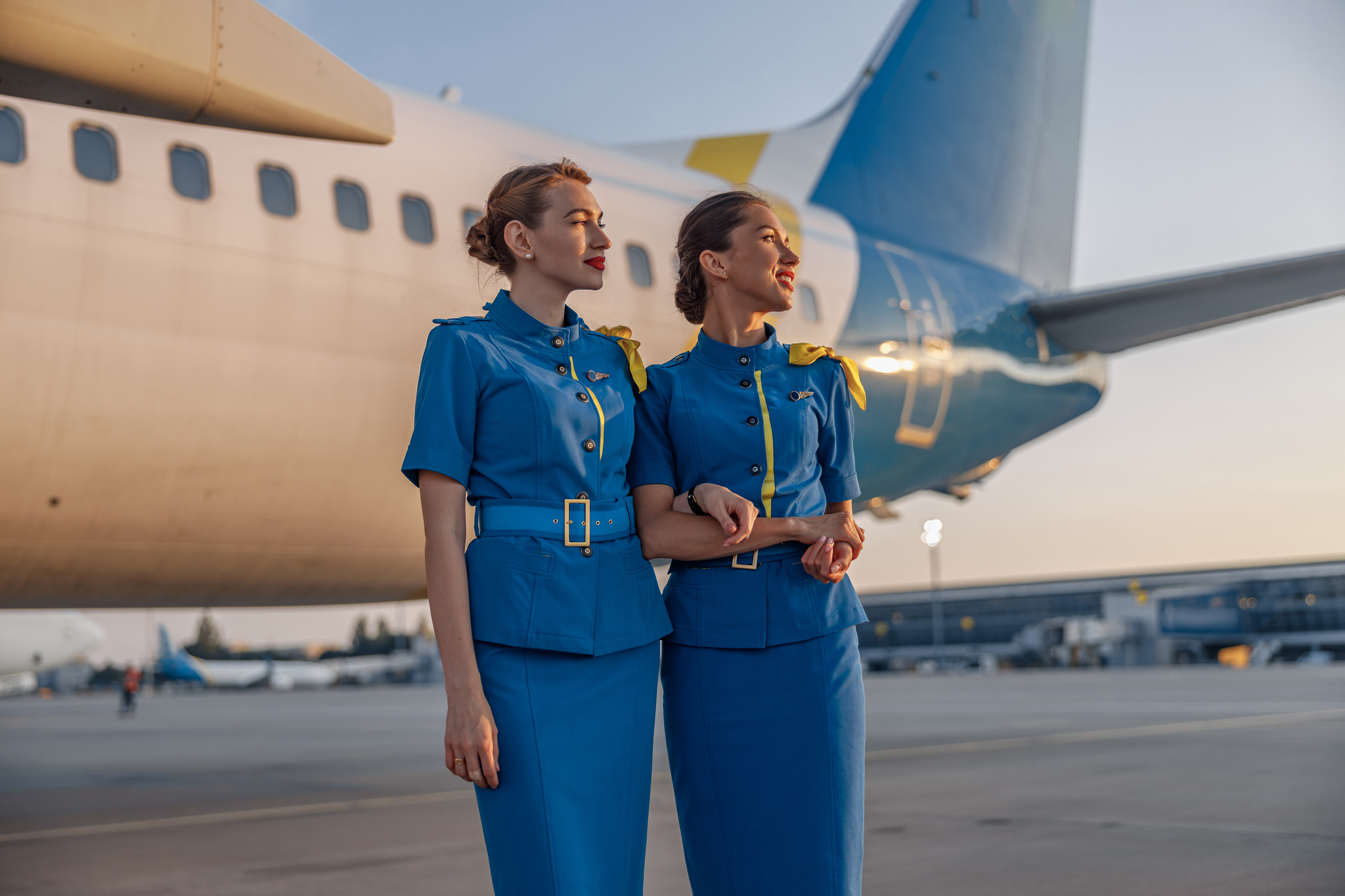 Two beautiful air hostesses in blue uniform smiling away, standing in front of a big passenger airplane in airport at sunset. | Source: Shutterstock