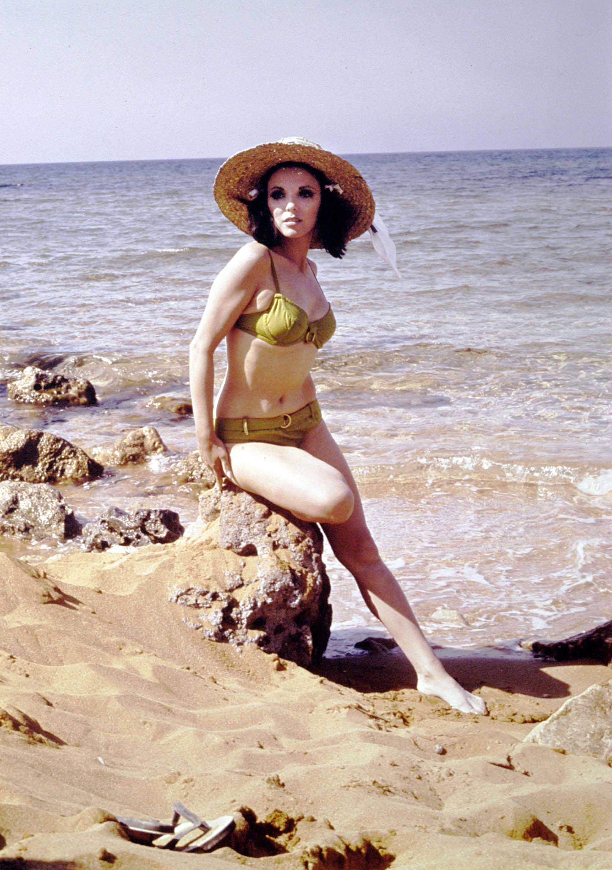 Joan Collins poses on a rocky beach in a moss green bikini with gold ring detailing. Wearing a wide straw sunhat with a white ribbon tied at the back, she gazes off-camera as waves lap the shore behind her. A pair of sandals rests in the foreground on the sand.