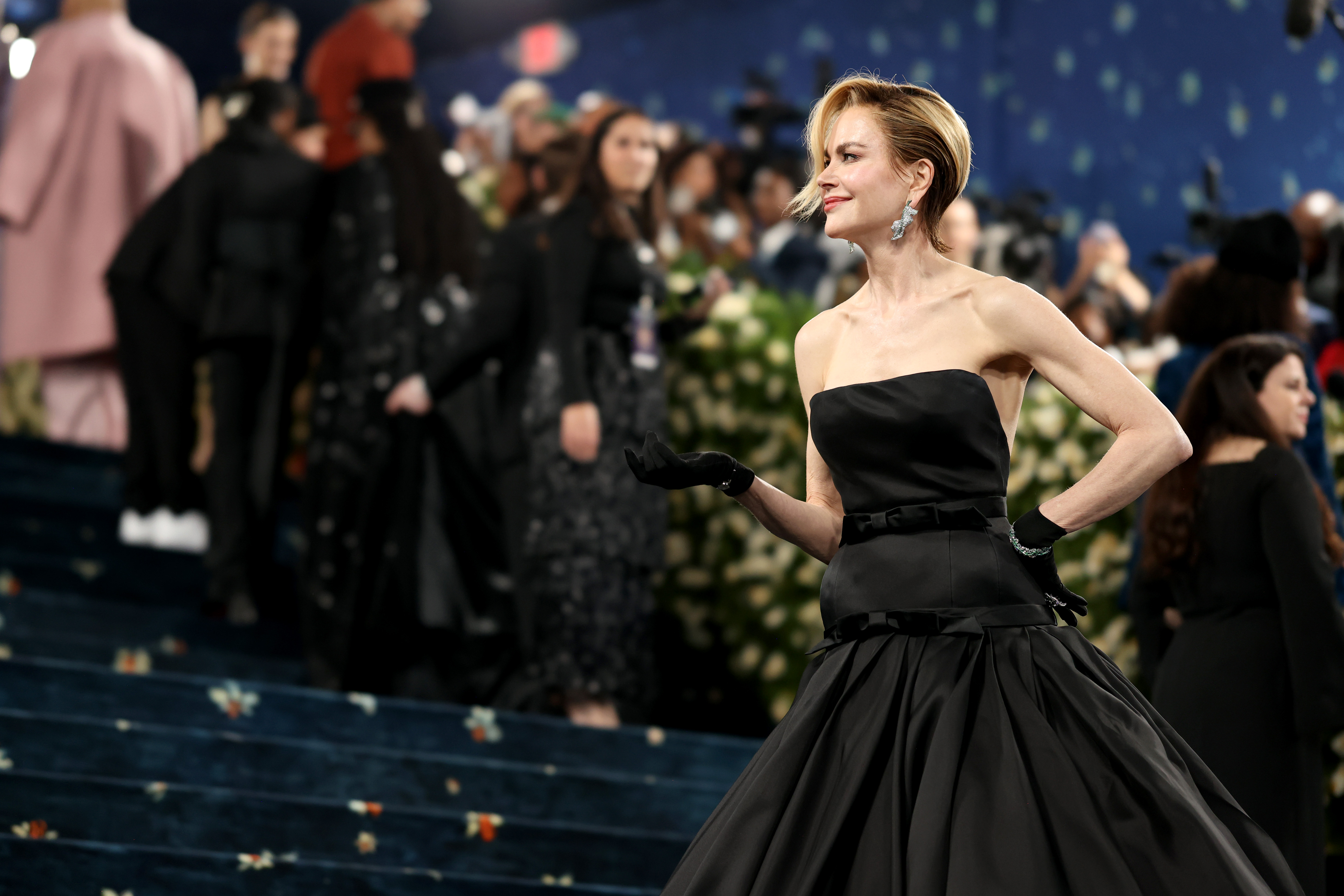 Nicole Kidman flashes a poised smile at the crowd as she poses on the red carpet, her arm resting gracefully on her waist. The gesture draws attention to a section of her gown, offering a glimpse of its textured fabric under the glow of camera flashes.
