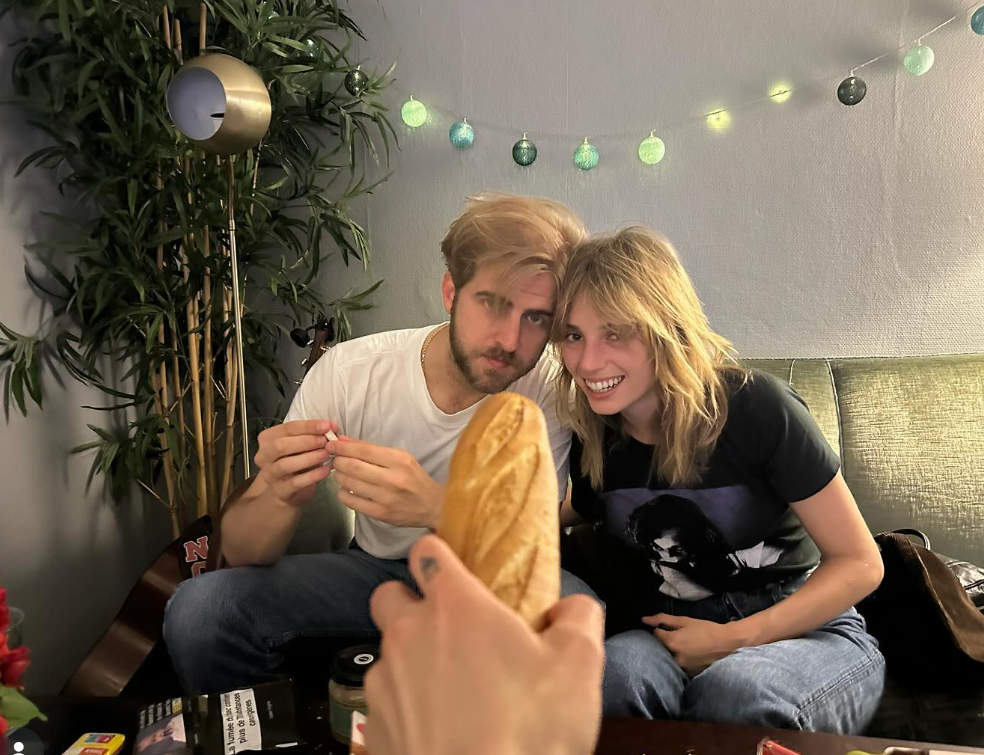 Maya Hawke and Christian Hutson sit close together on a couch at home, smiling warmly beneath soft string lights and greenery. | Source: Instagram/christianleehutson