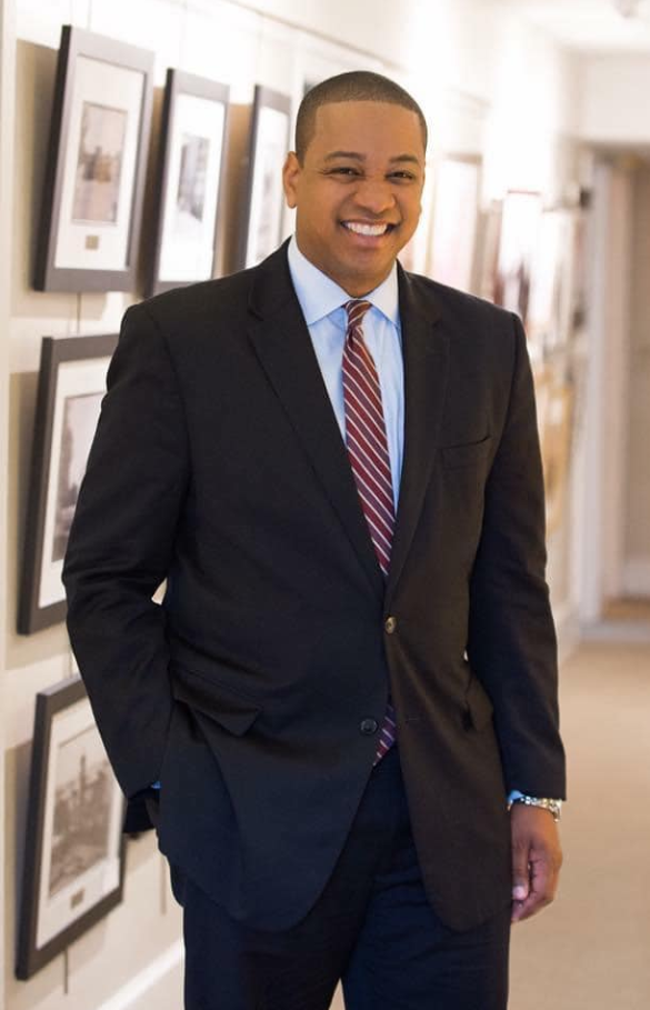 Justin Fairfax stands in a hallway lined with framed photos, dressed in a dark suit and striped tie.. | Source: Facebook/JustinFairfax