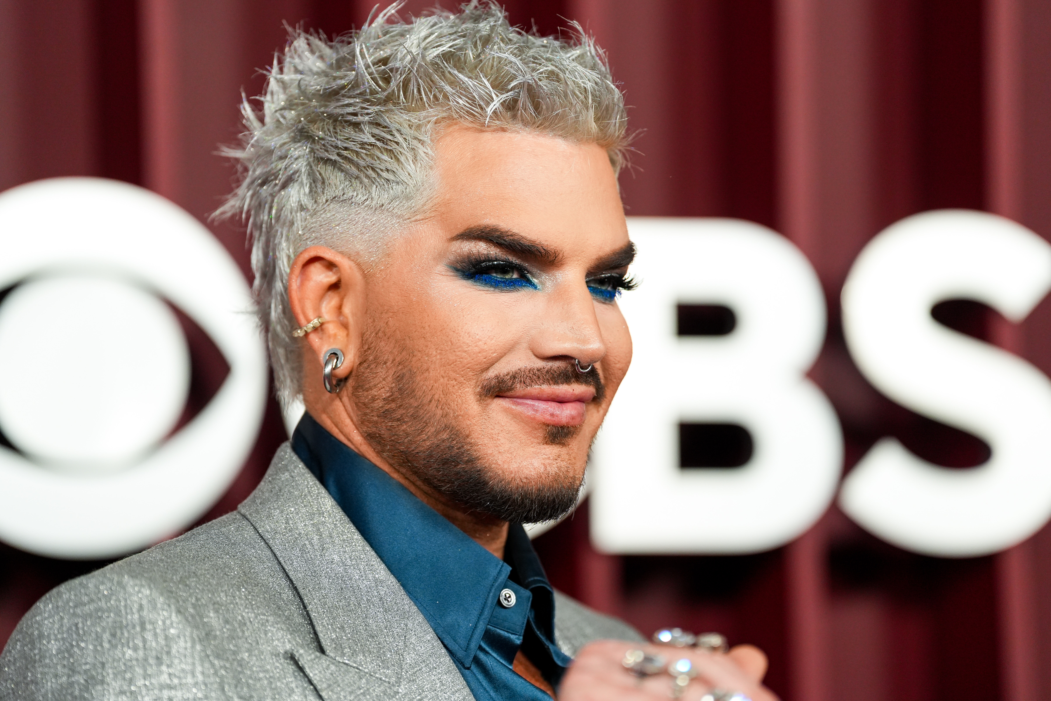 Adam Lambert flashes a knowing smile as his platinum-textured hairstyle and shimmering suit reflect the lights. The deep navy shirt and vivid blue eyeshadow complete a look that is equal parts polished sophistication and artistic boldness.