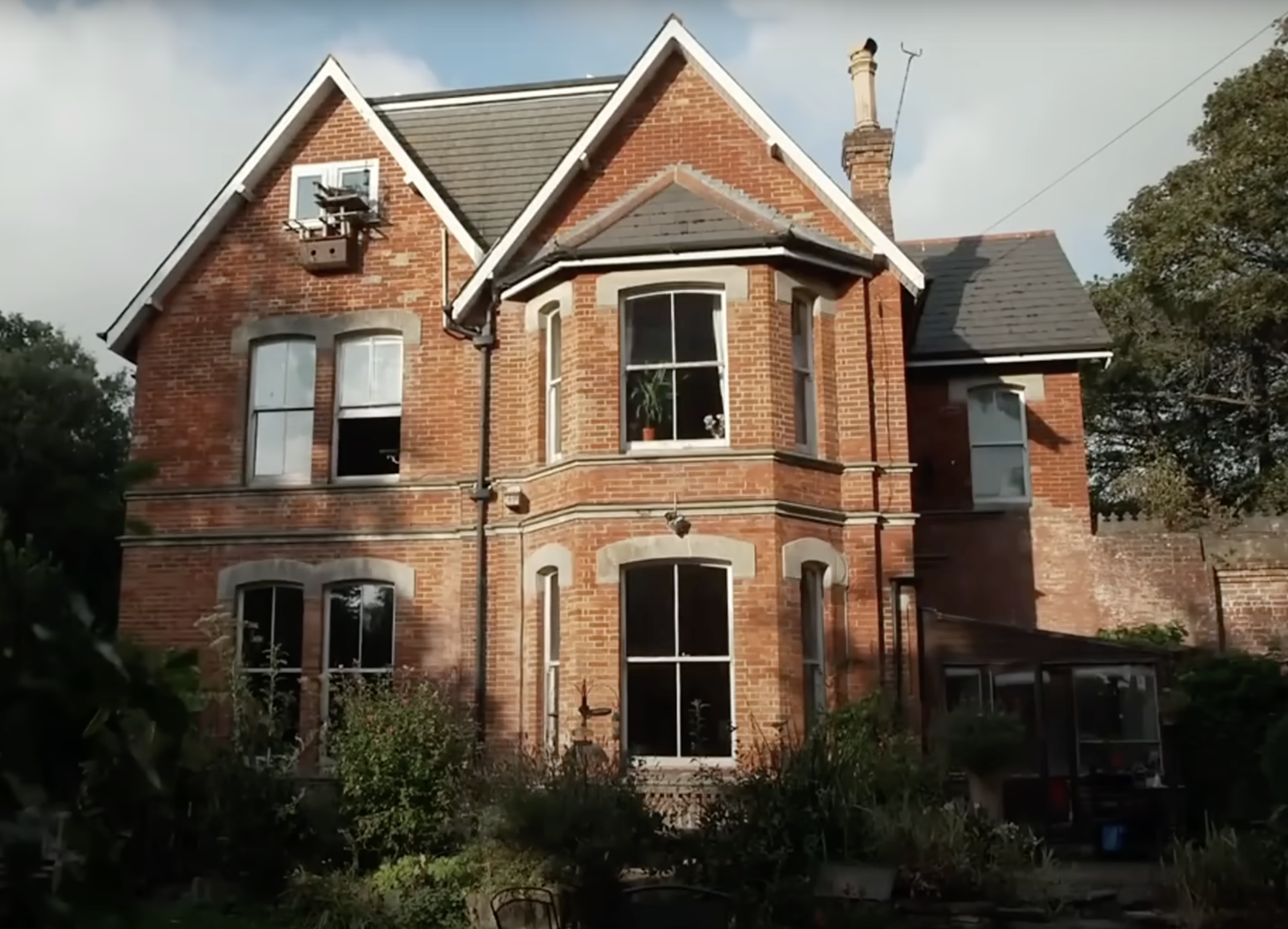 The exterior of Jane Goodall's red-brick childhood home in Bournemouth, England, is shown in daylight. Several large windows overlook the garden, where plants grow wild near the base of the house. | Source: YouTube/@CBSSundayMorning