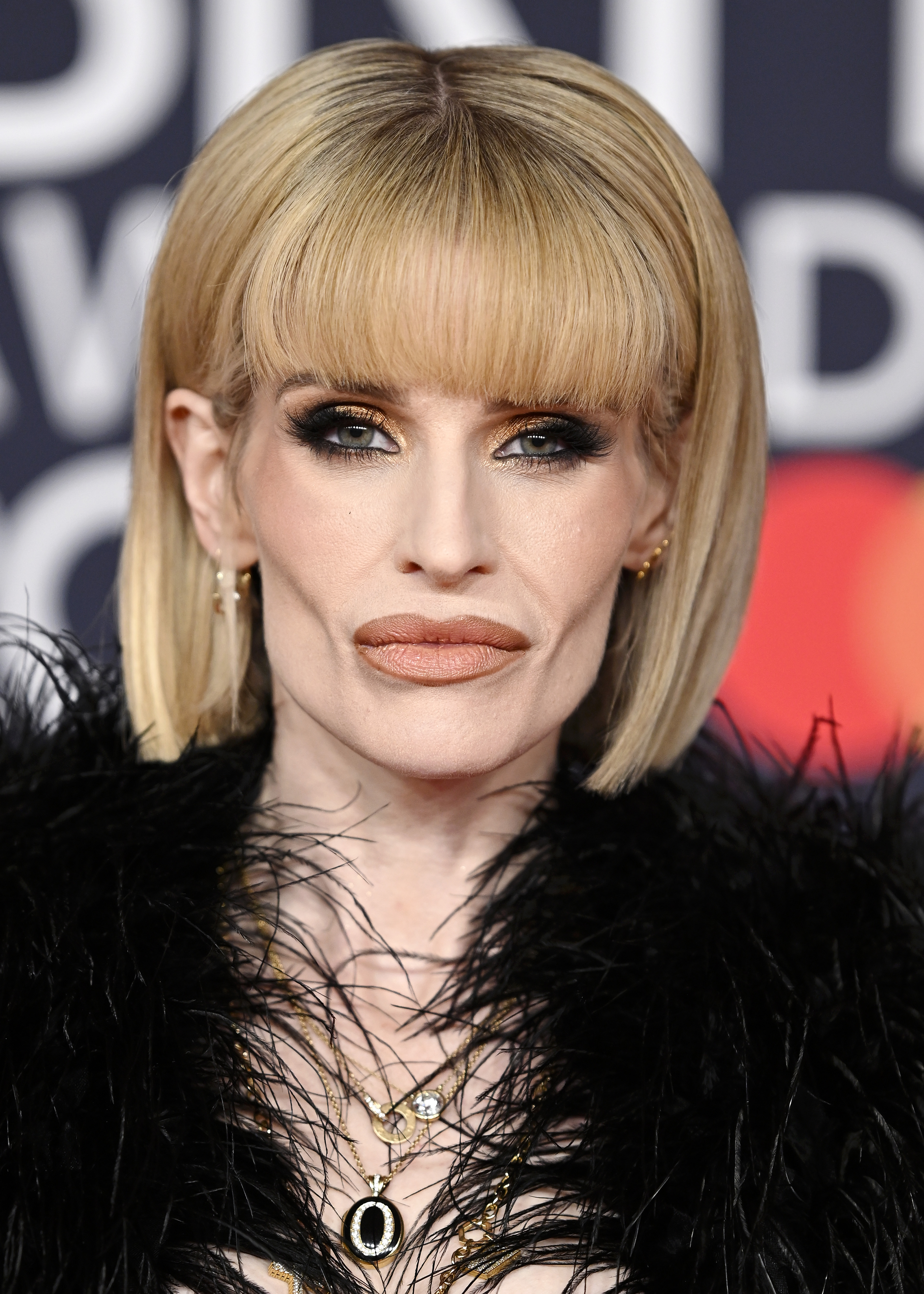 In February, Kelly Osbourne showcases striking sophistication with a sleek blonde bob, dramatic smoky eye makeup, and layered gold jewelry. Framed by textured black feathers, the look blends old-Hollywood glamour with the edgy style that has long defined her public image.